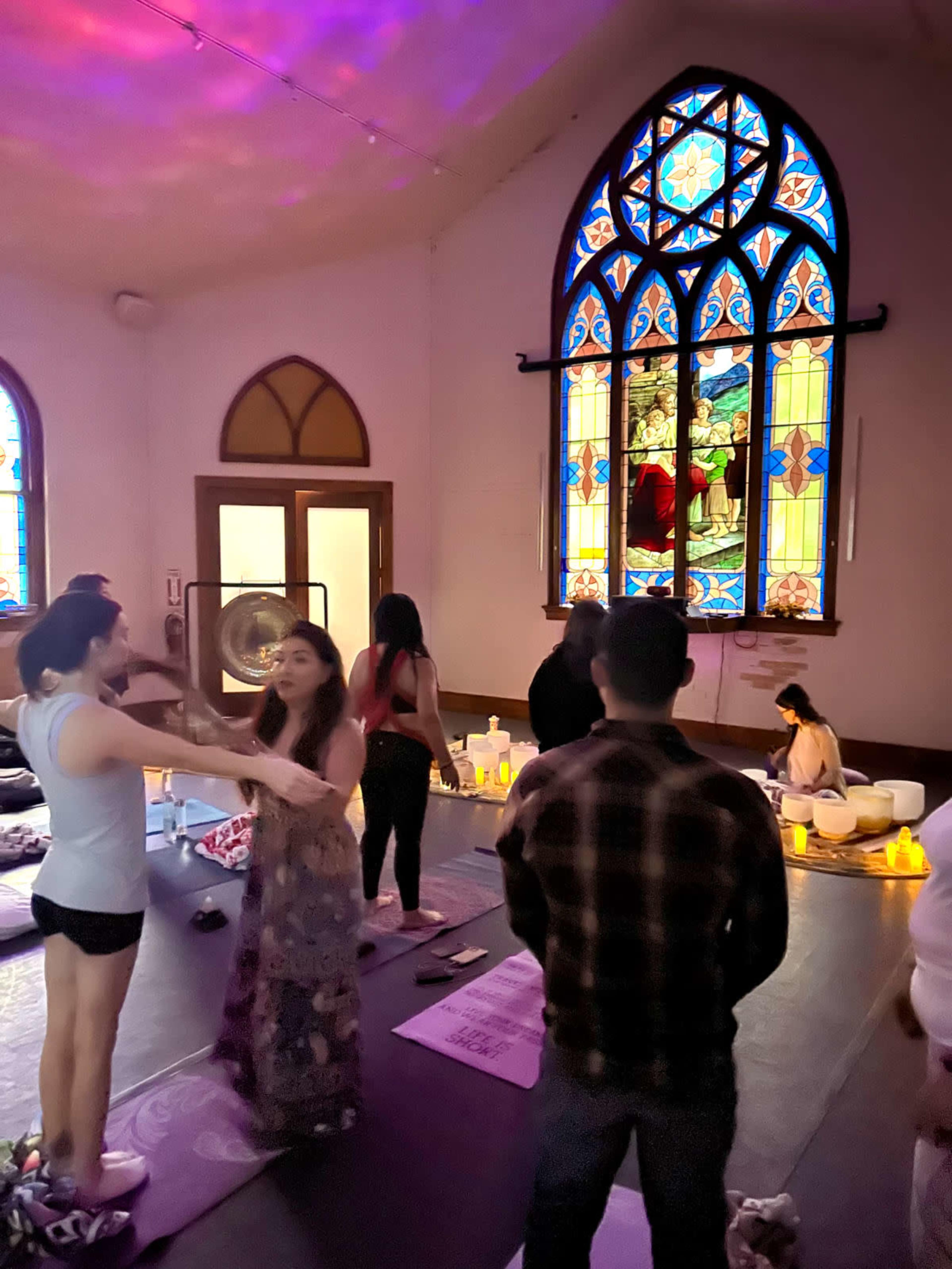 A group of individuals participates in a yoga session inside a room with stained glass windows, surrounded by candles and various yoga mats.