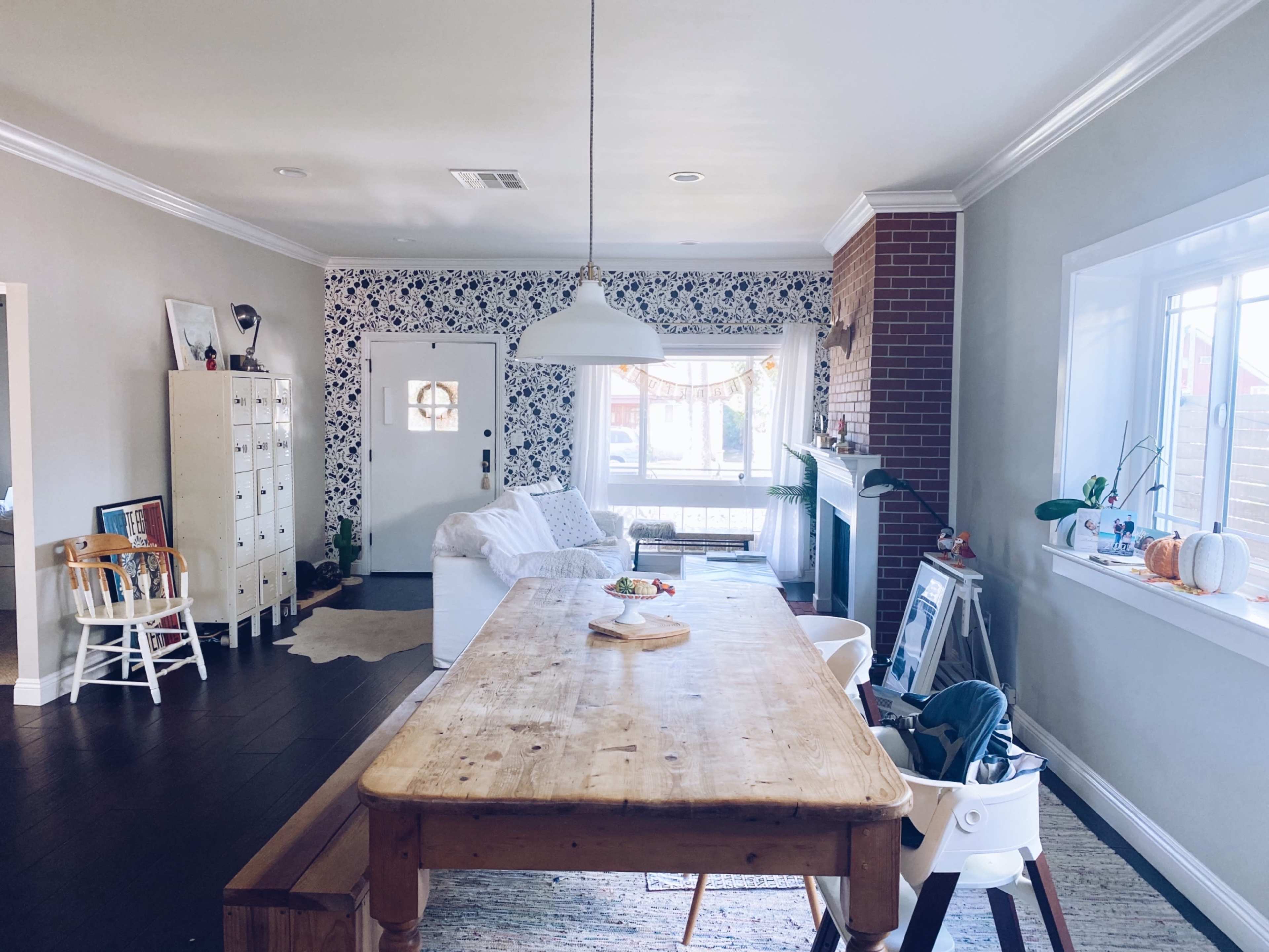A bright living room with a white sofa, a wooden dining table, and a brick fireplace, contrasting with patterned wallpaper.