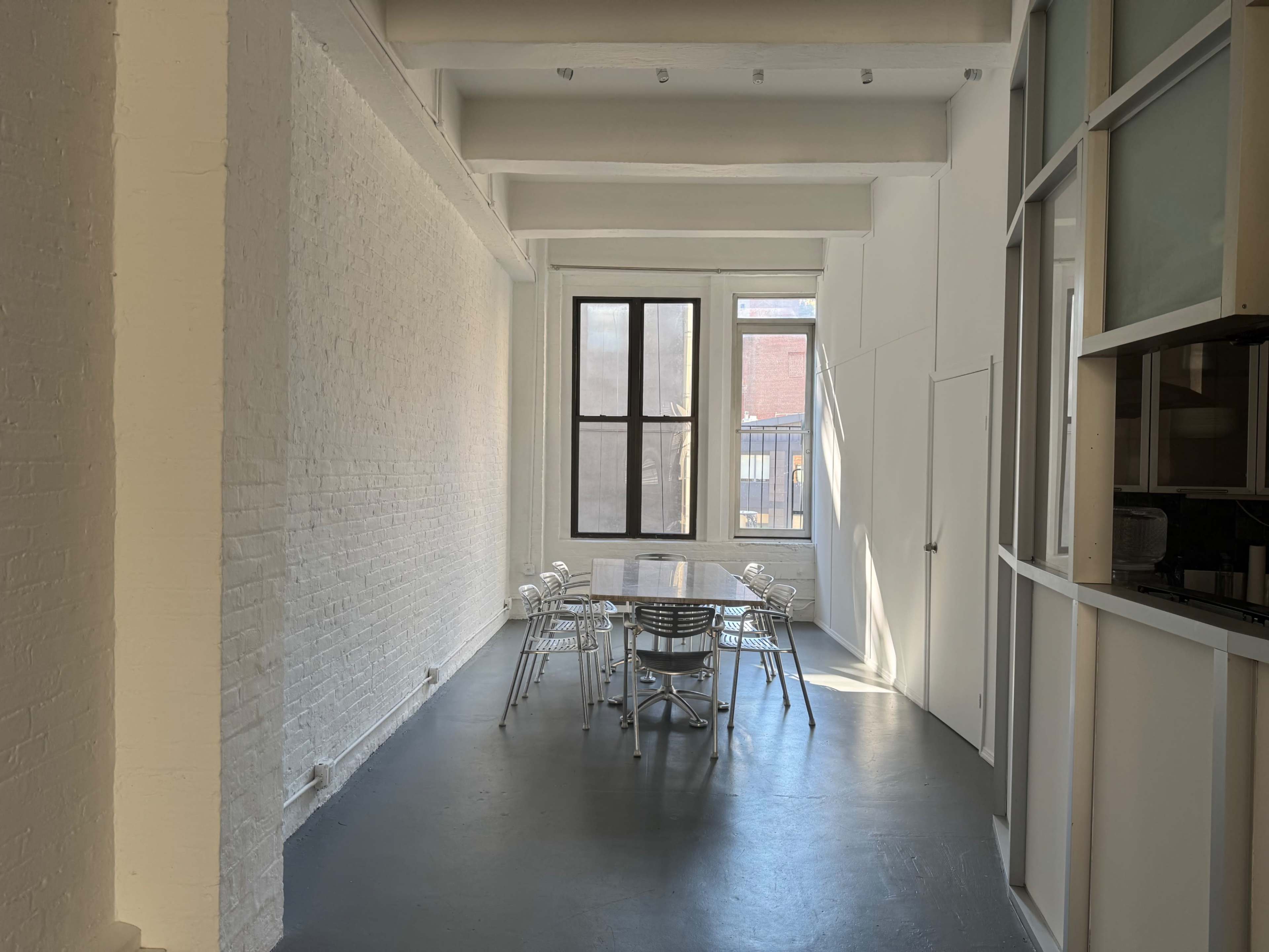 A modern, minimalistic dining area features a long table surrounded by several chairs, illuminated by natural light from large windows.