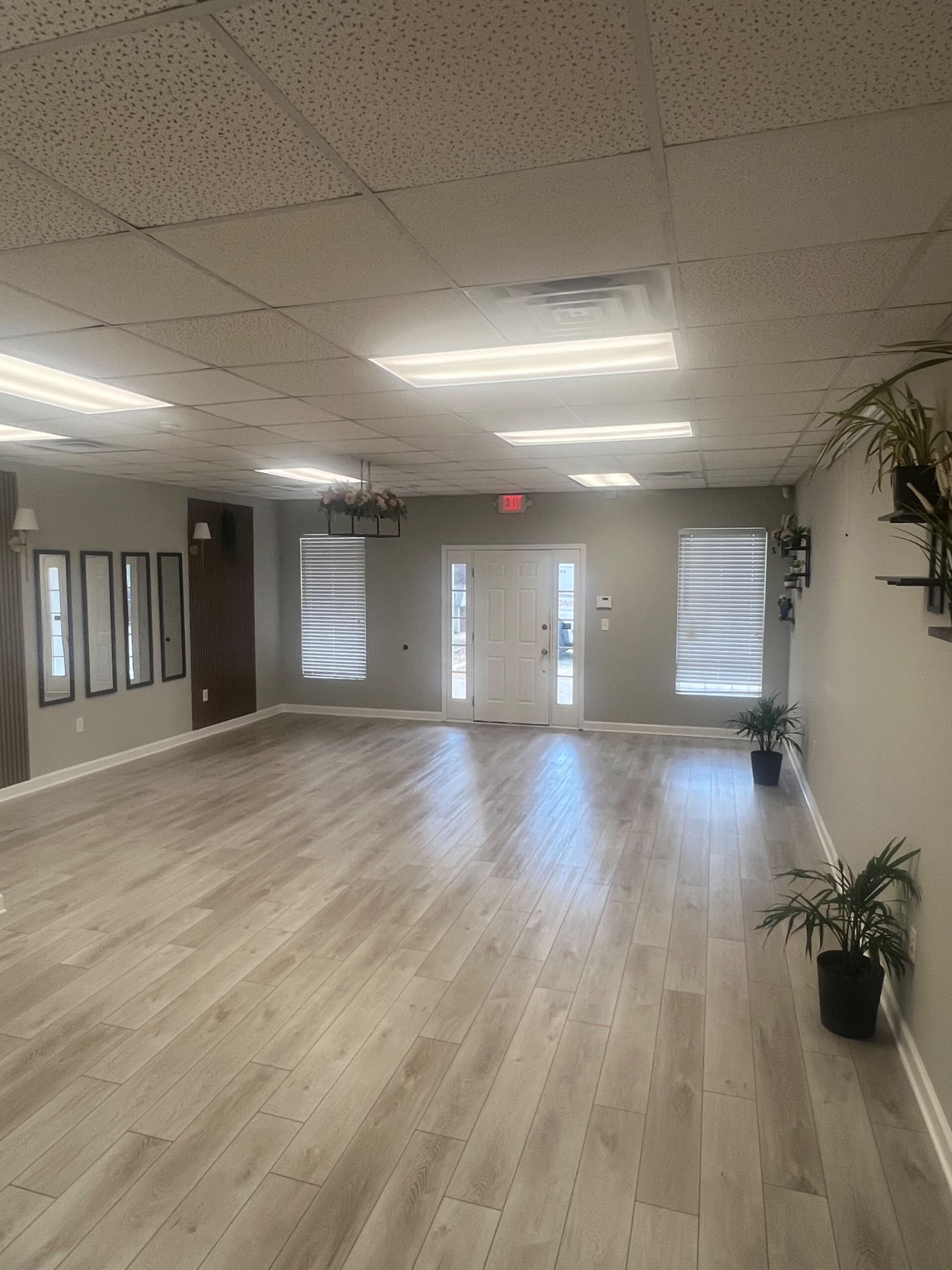 The image shows an empty room with light wood flooring, gray walls, plants in the corners, and a door at the end leading outside.