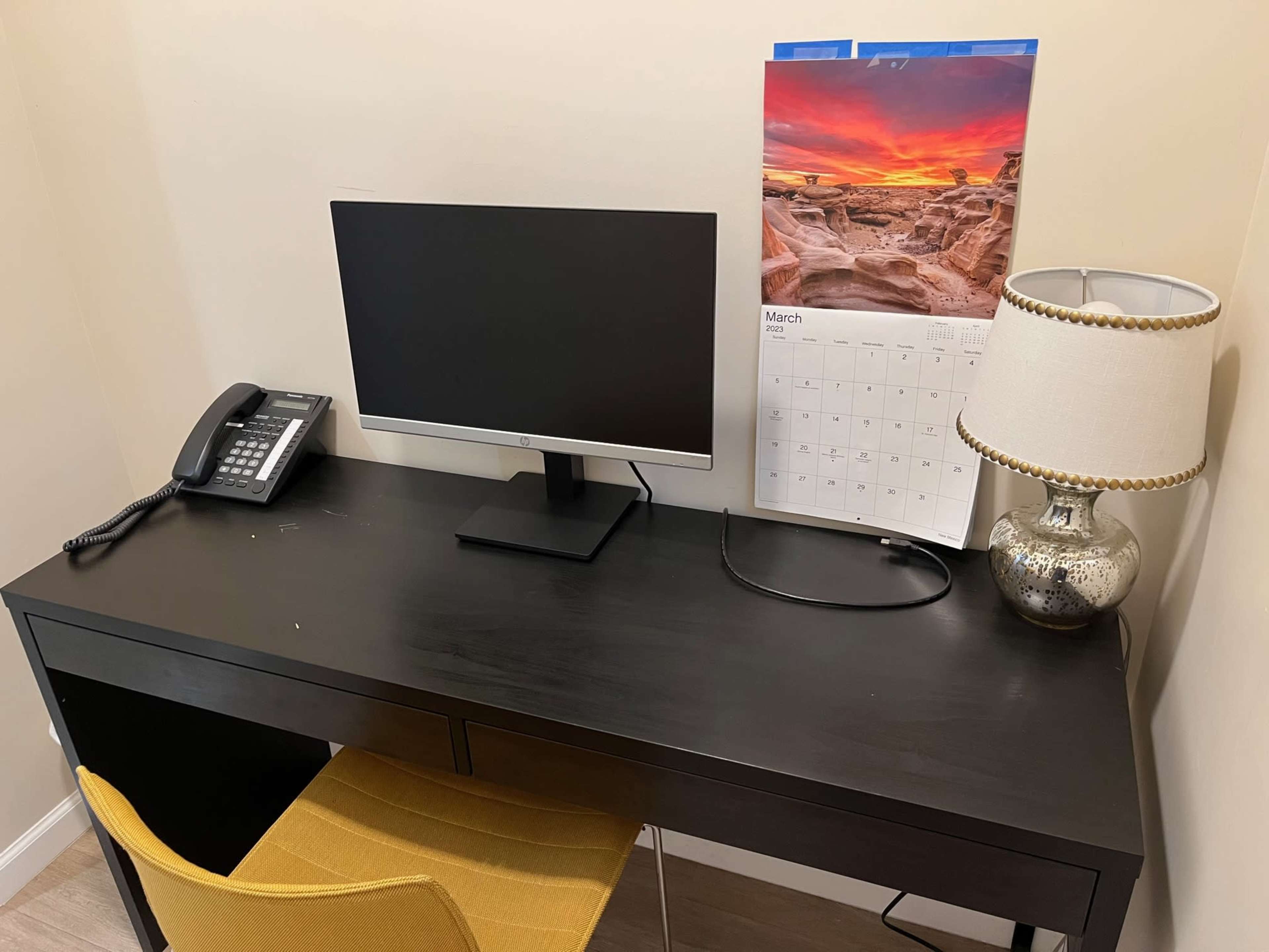 A black desk with a computer monitor, a telephone, a lamp, and a calendar is set against a beige wall.