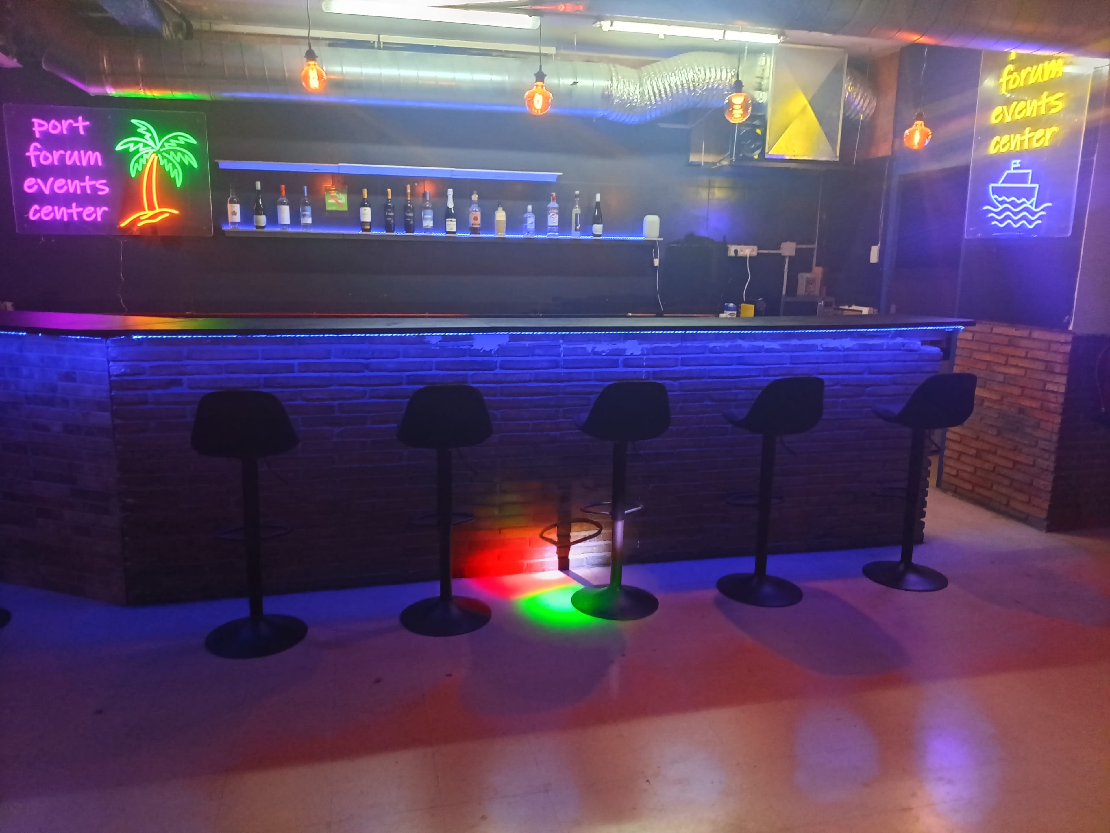 The image shows a bar area with illuminated bottles on a shelf, surrounded by colorful neon signs and empty black stools.