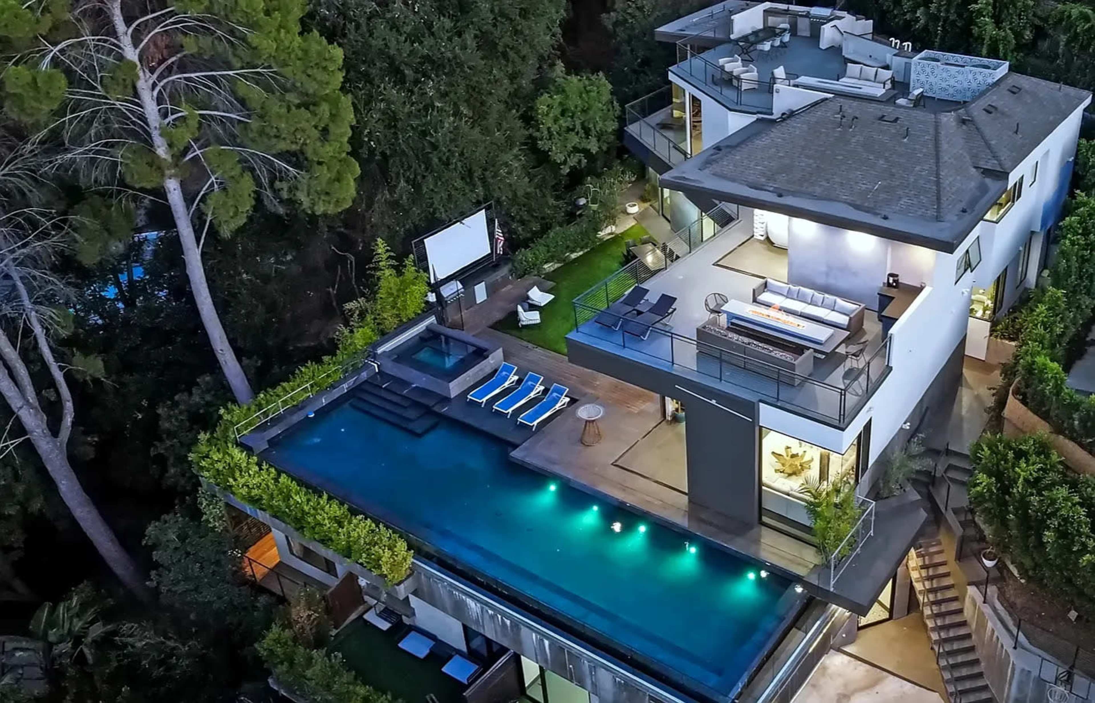 The image shows a modern multi-story house with a large rooftop terrace and an infinity pool surrounded by landscaping and lounge chairs.