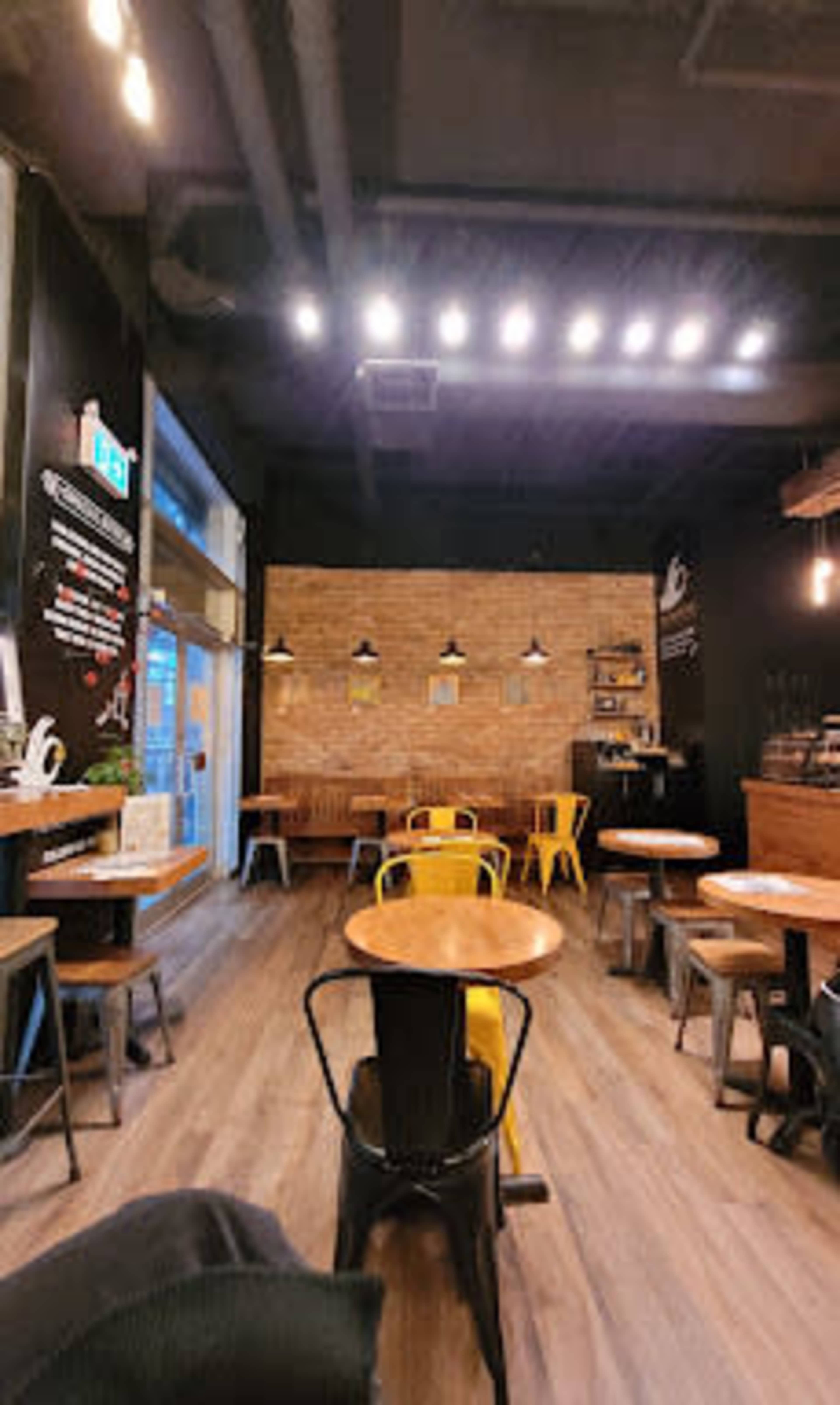 The image shows a cozy café interior with wooden tables, yellow and black chairs, and exposed brick walls.