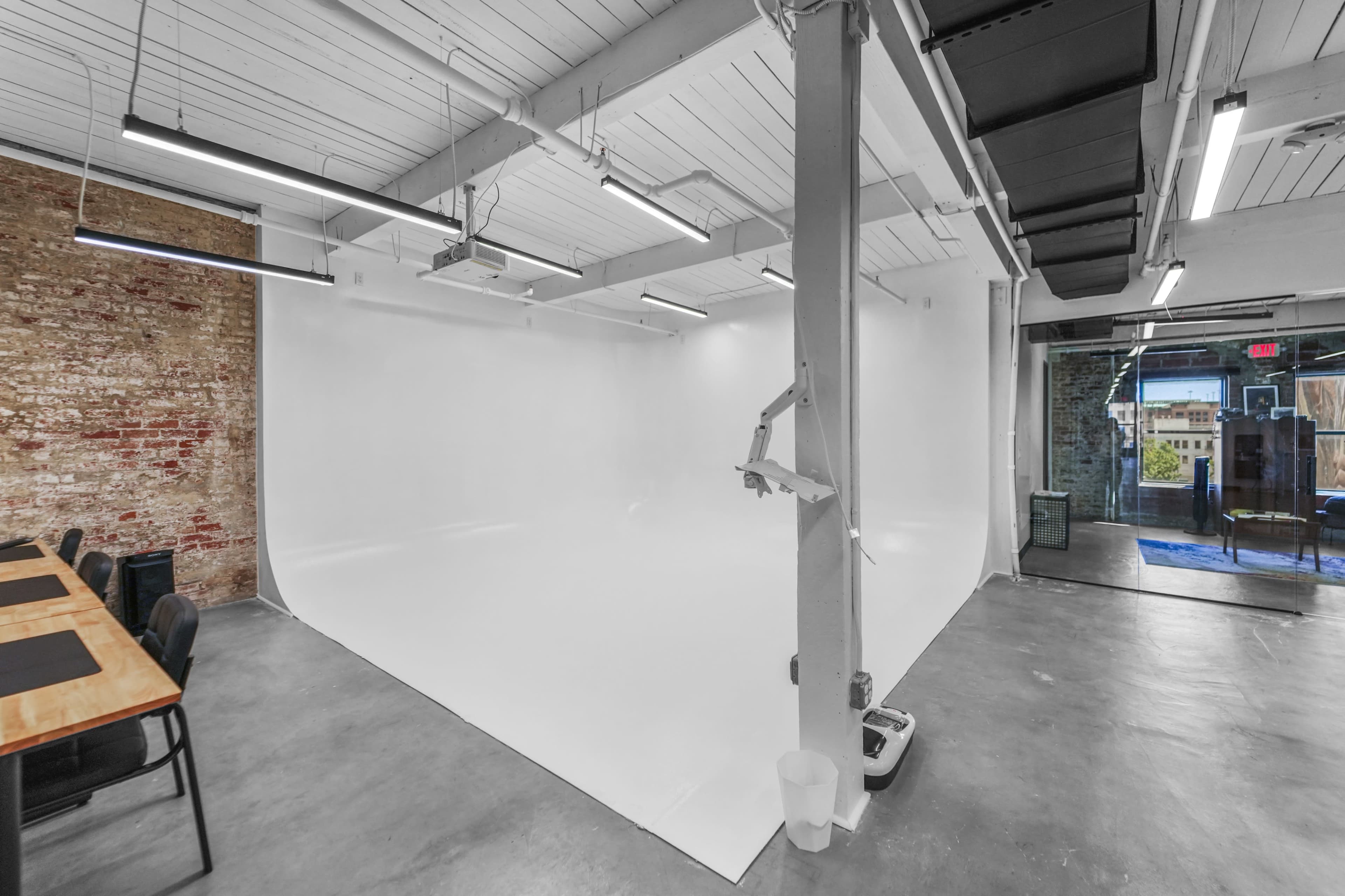 The image shows a spacious studio with a white backdrop, concrete flooring, and exposed brick walls, featuring a long table and chairs on one side.