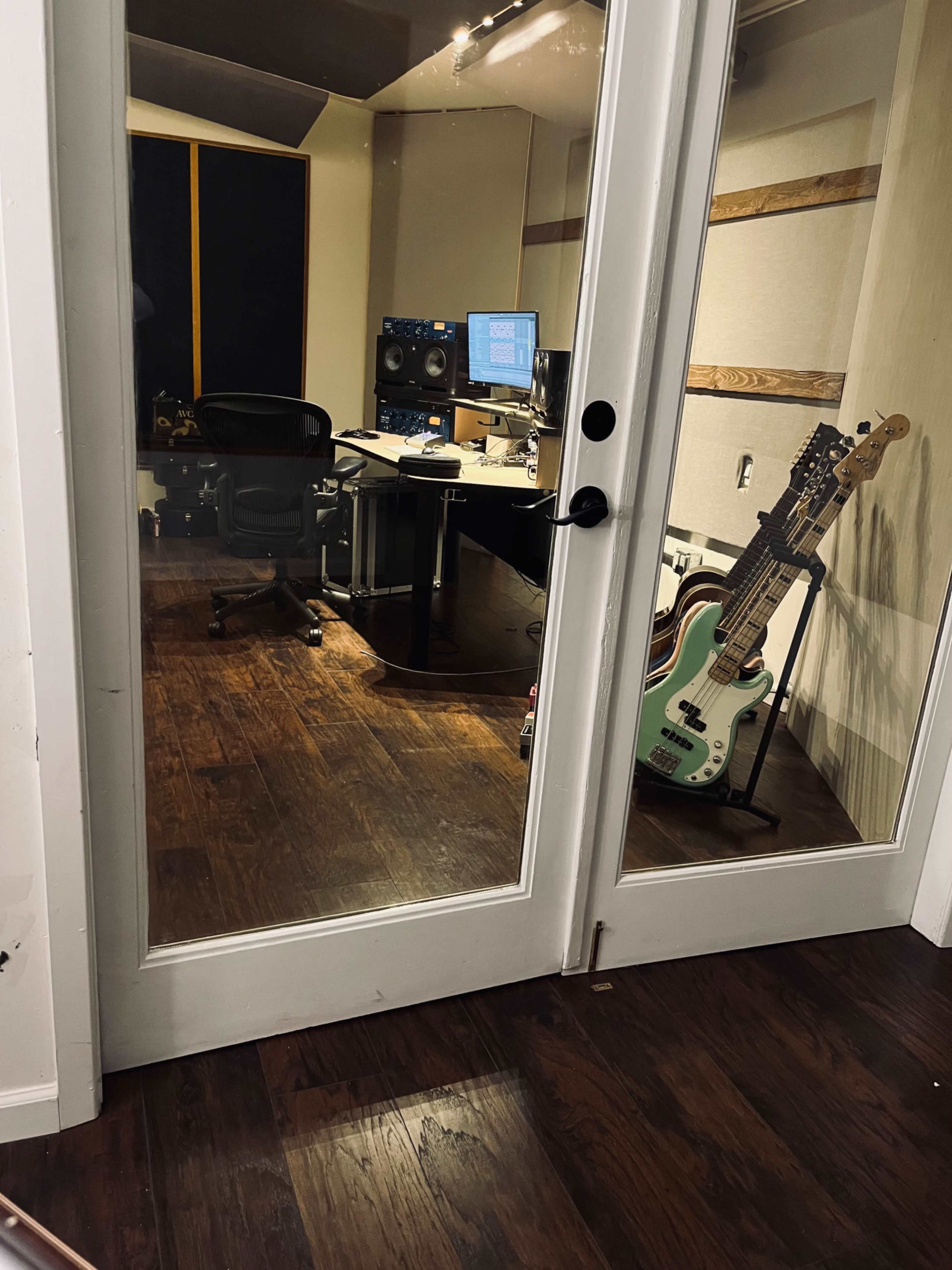 A music studio is visible through glass doors, featuring a desk with computers, audio equipment, and a pair of guitars leaning against the wall.