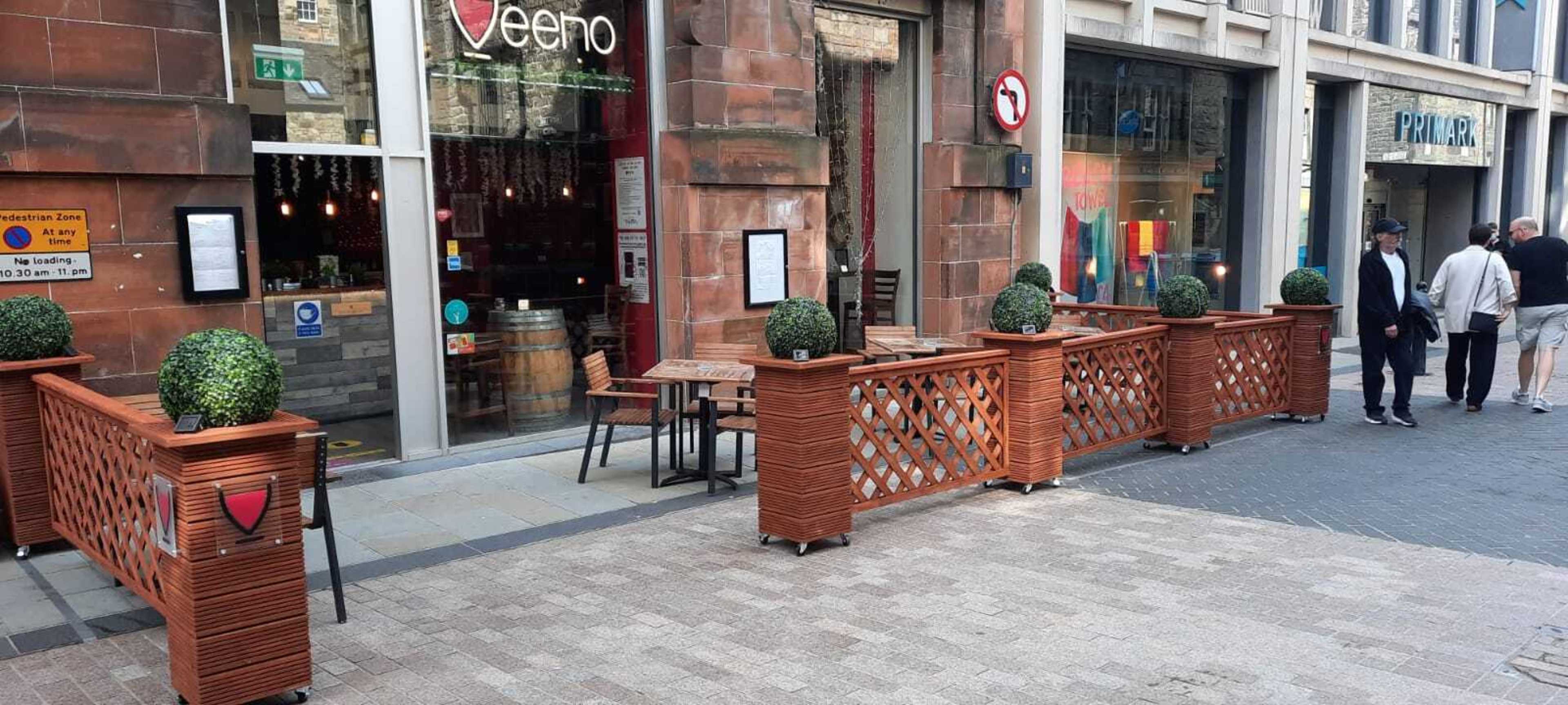The image shows an outdoor seating area with wooden barriers and potted topiaries in front of a restaurant called "Veenoo" on a street with pedestrians walking by.