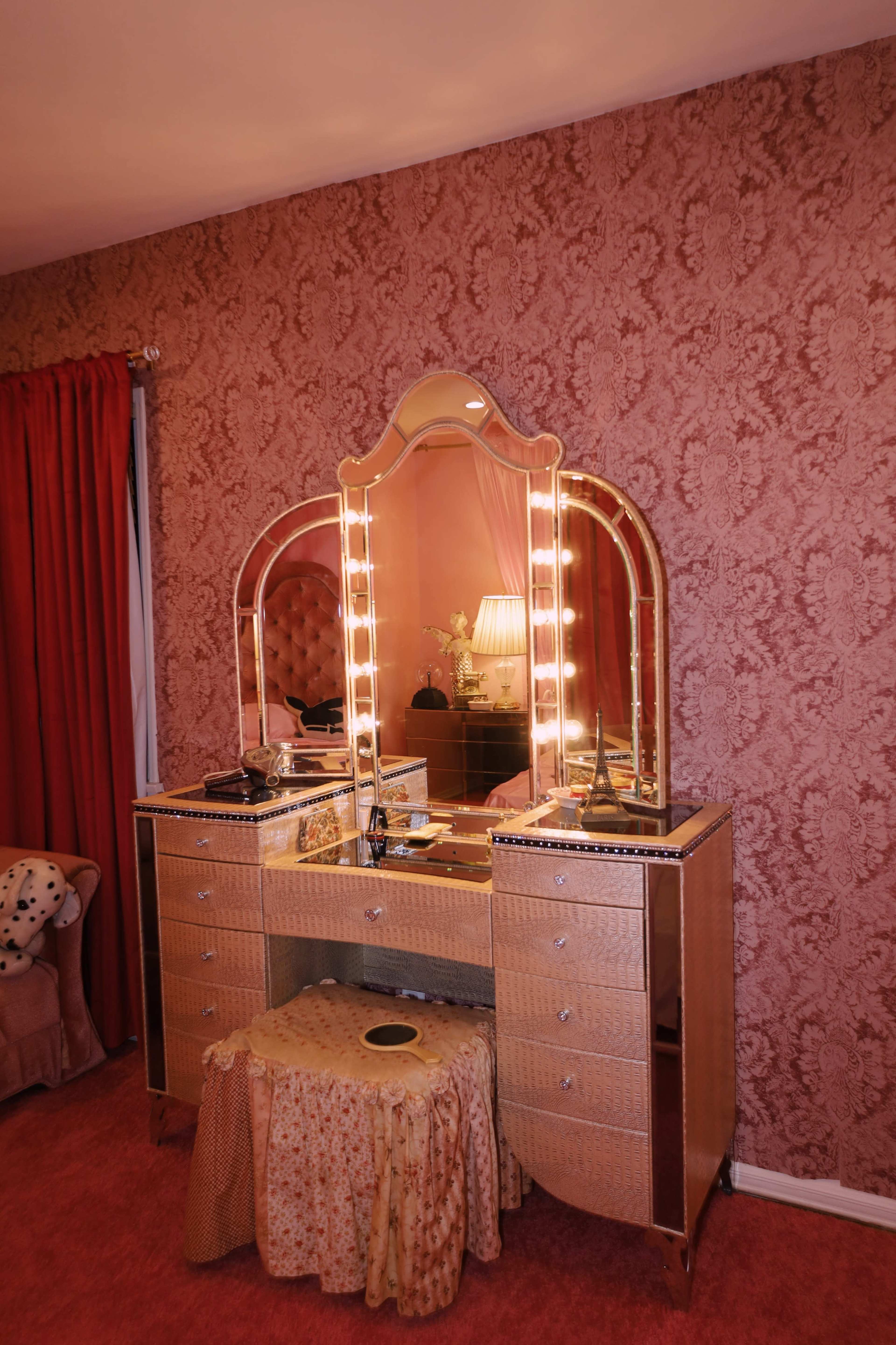 A vintage vanity set with a large mirror and lights is positioned against a patterned wallpaper in a room featuring red curtains and carpet.