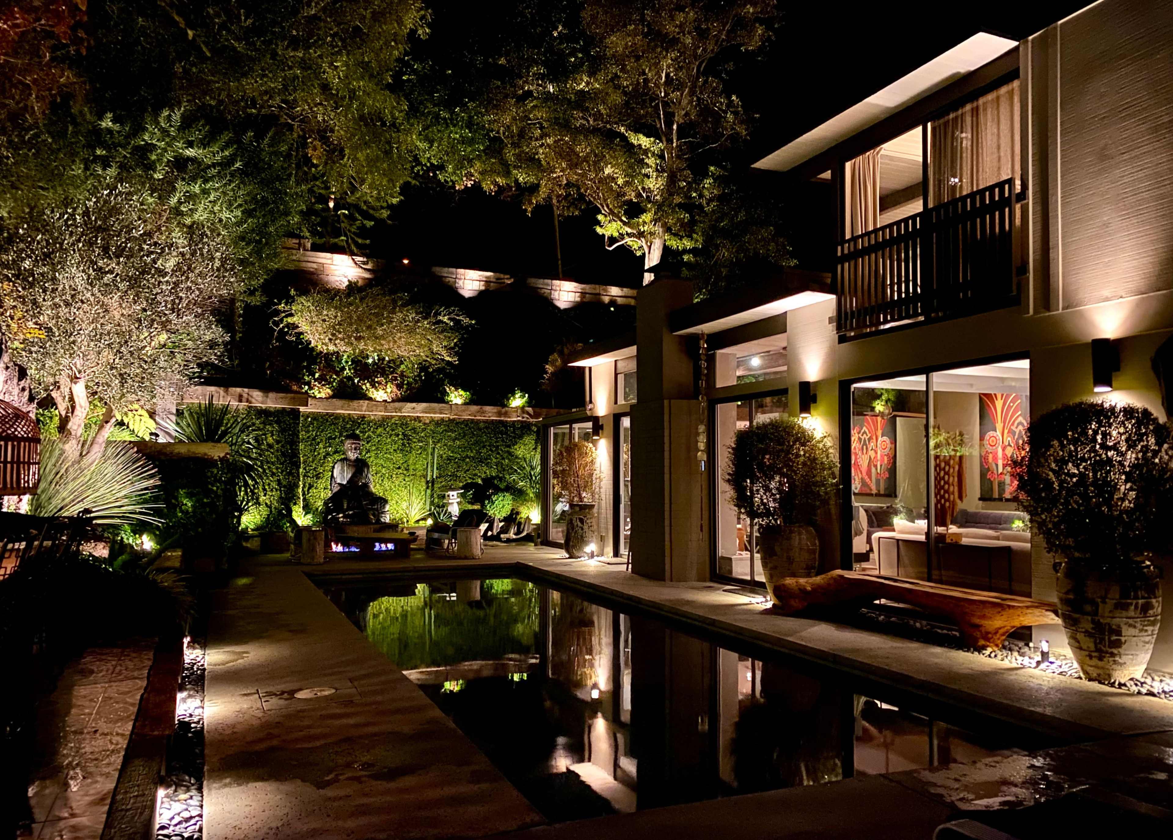 A modern outdoor pool area at night, featuring landscaped greenery, illuminated trees, and a statue surrounded by decorative elements.