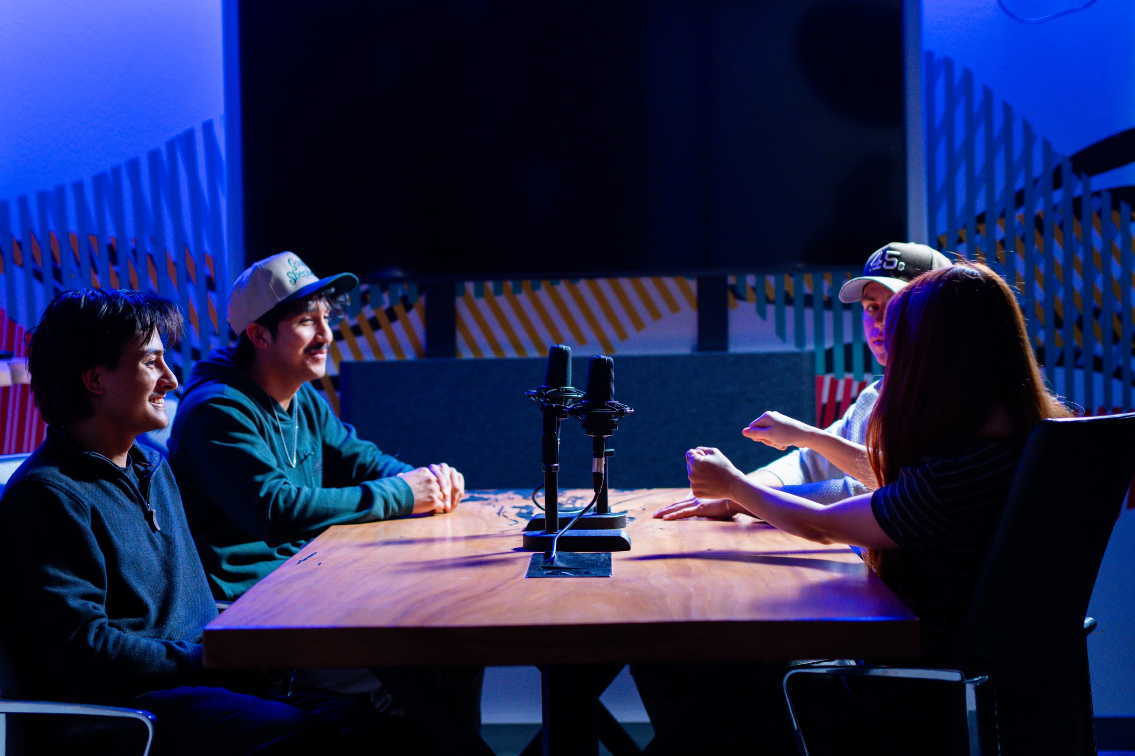 Four people sit around a wooden table with microphones in a brightly colored studio setting.
