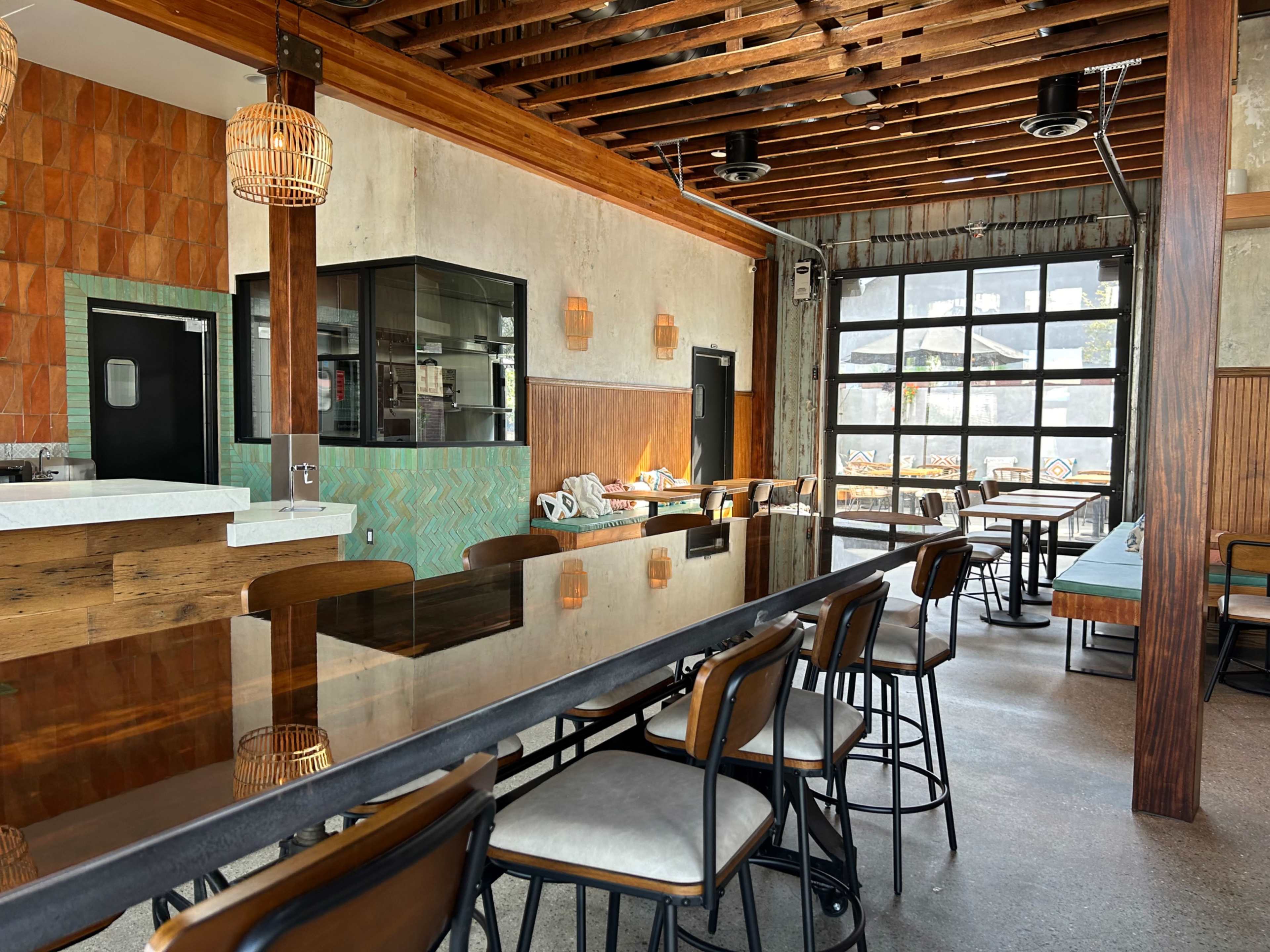 A modern restaurant interior with wooden beams, a long bar counter, and tables arranged near large windows.