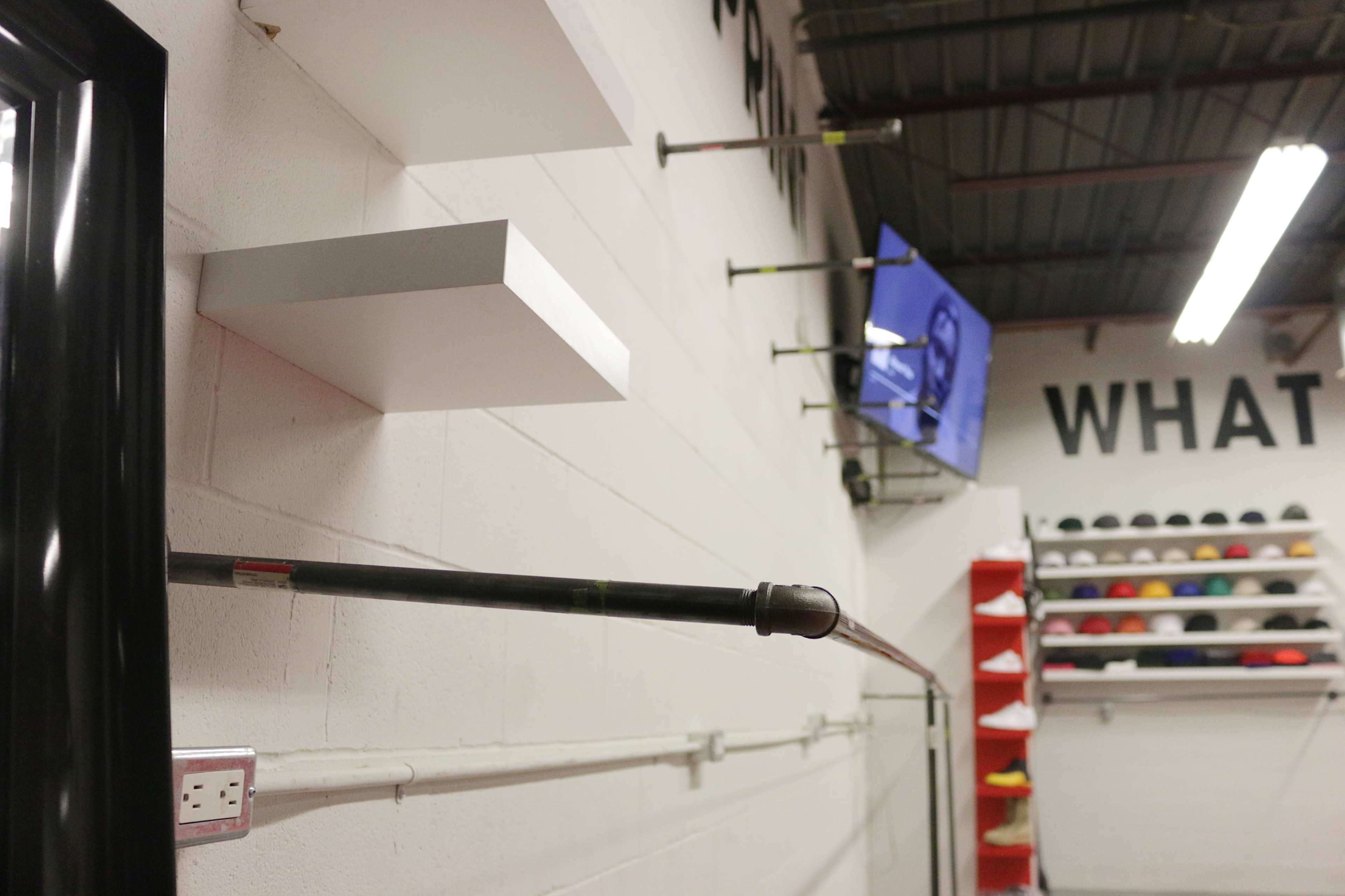 A wall with two white shelves, a horizontal pipe, an electrical outlet, and a television mounted above a row of colorful hats.