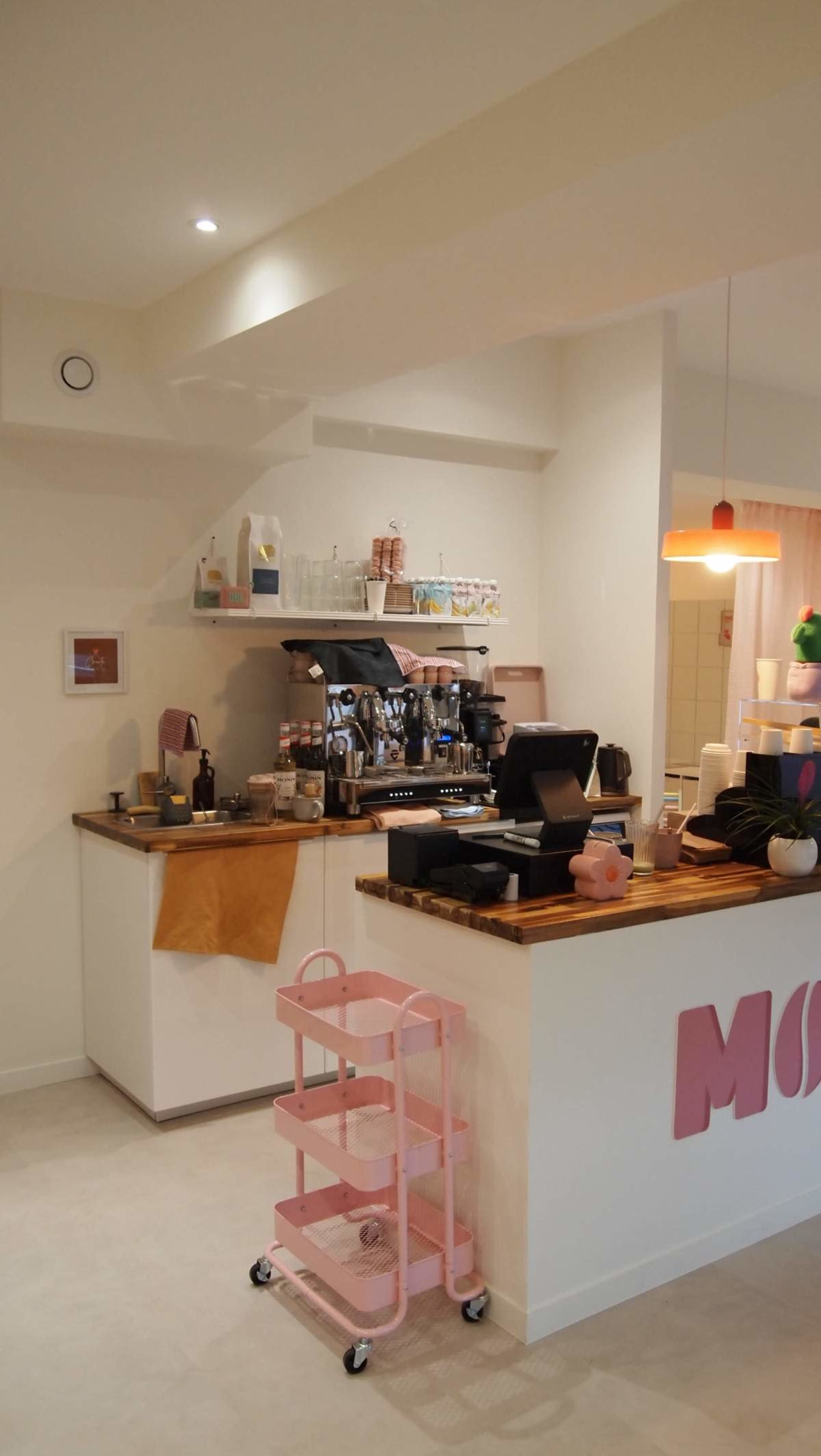 The image shows a well-organized coffee shop interior with a barista station, a wooden countertop, and a pastel pink utility cart.