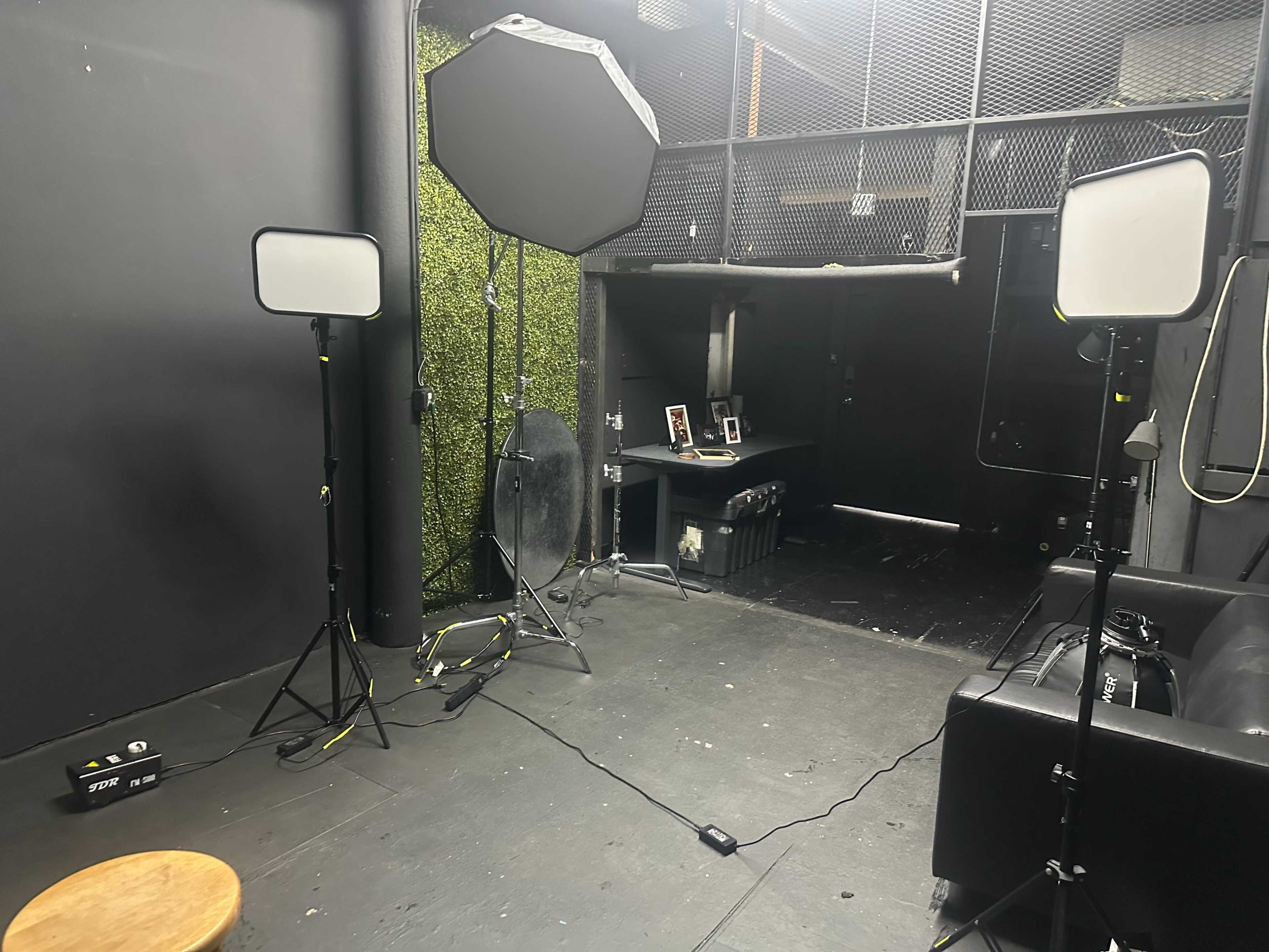 The image shows a studio setup with two light stands holding softboxes, a stool, and a black couch, all positioned near a dark backdrop and an open doorway leading to another room.