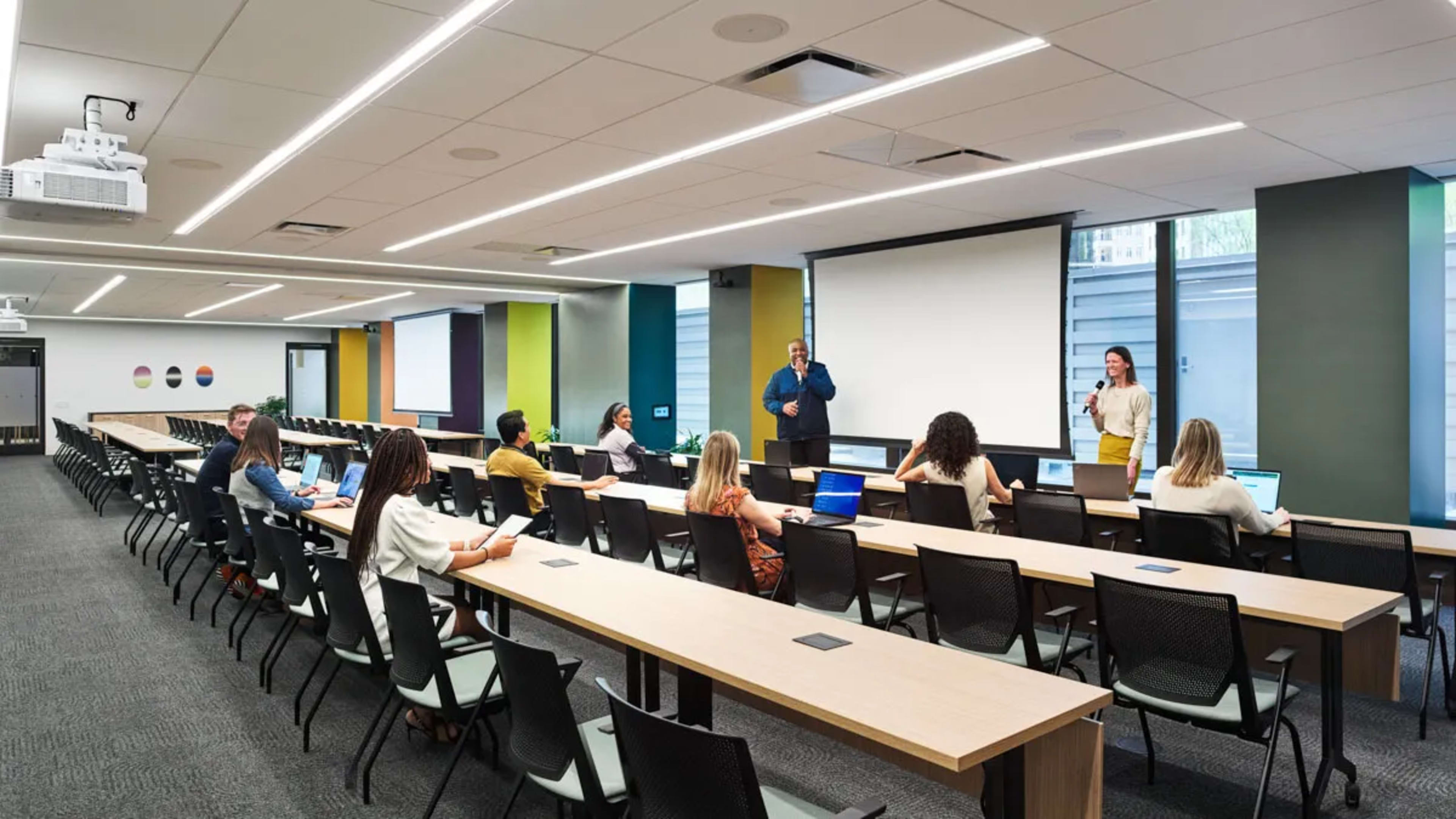 A spacious conference room is set up with long tables facing a presentation area, where two speakers are addressing an audience of seated participants using laptops.