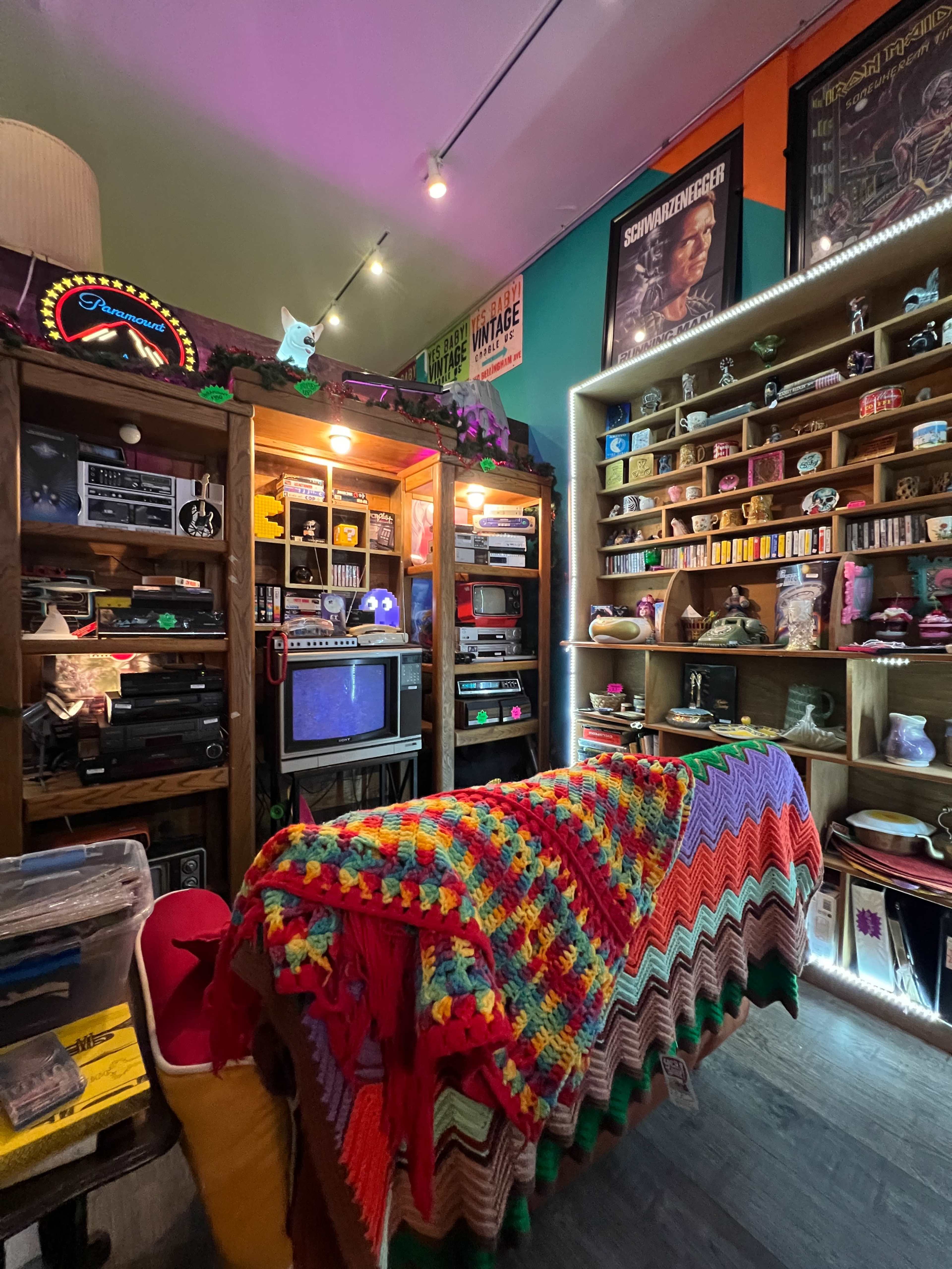 The image shows a vintage-themed room filled with various retro items, shelves of collectibles, a small television, and a colorful crocheted blanket draped over a chair.