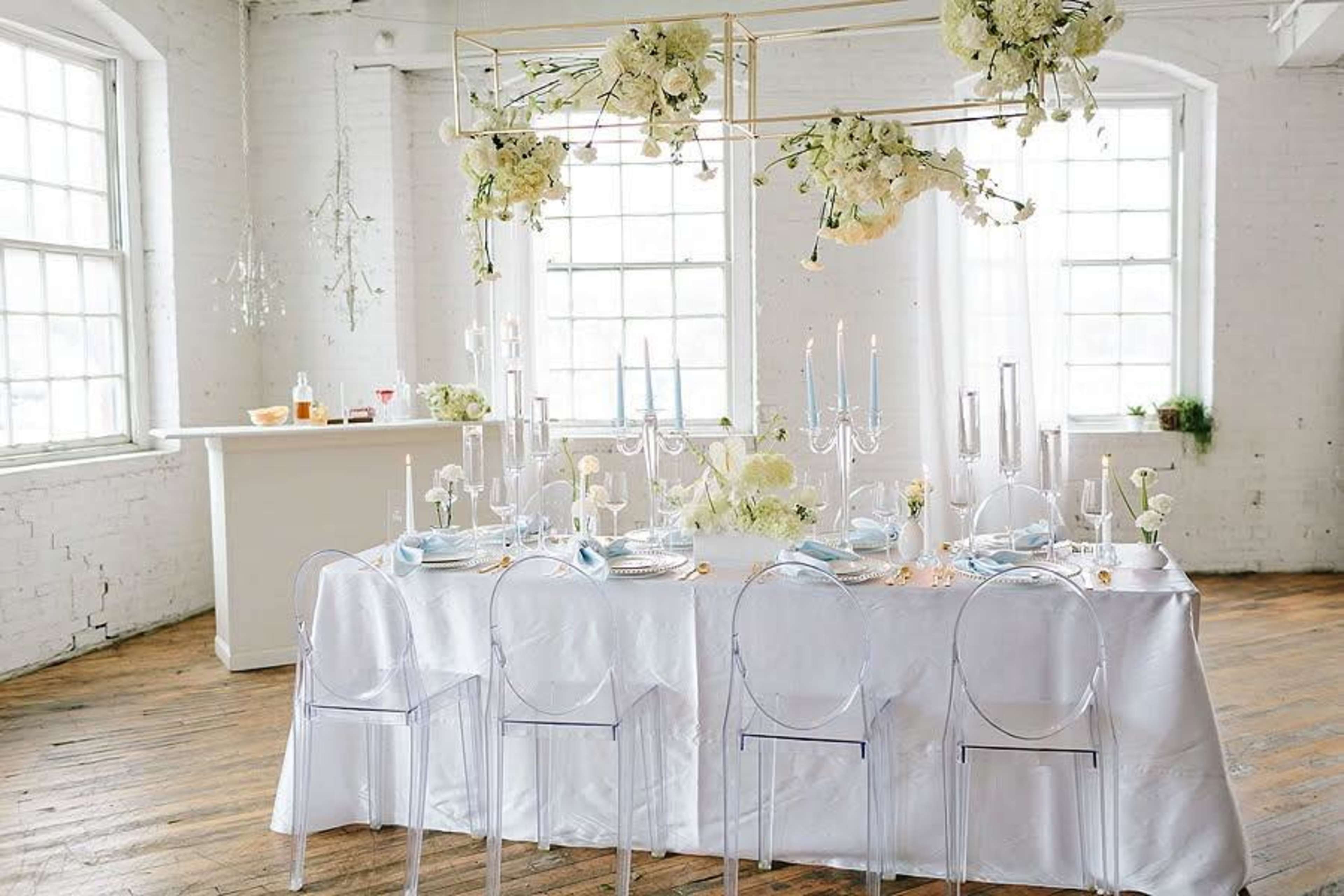 A dining table is elegantly set with floral arrangements, candles, and tableware in a bright, airy room with large windows.