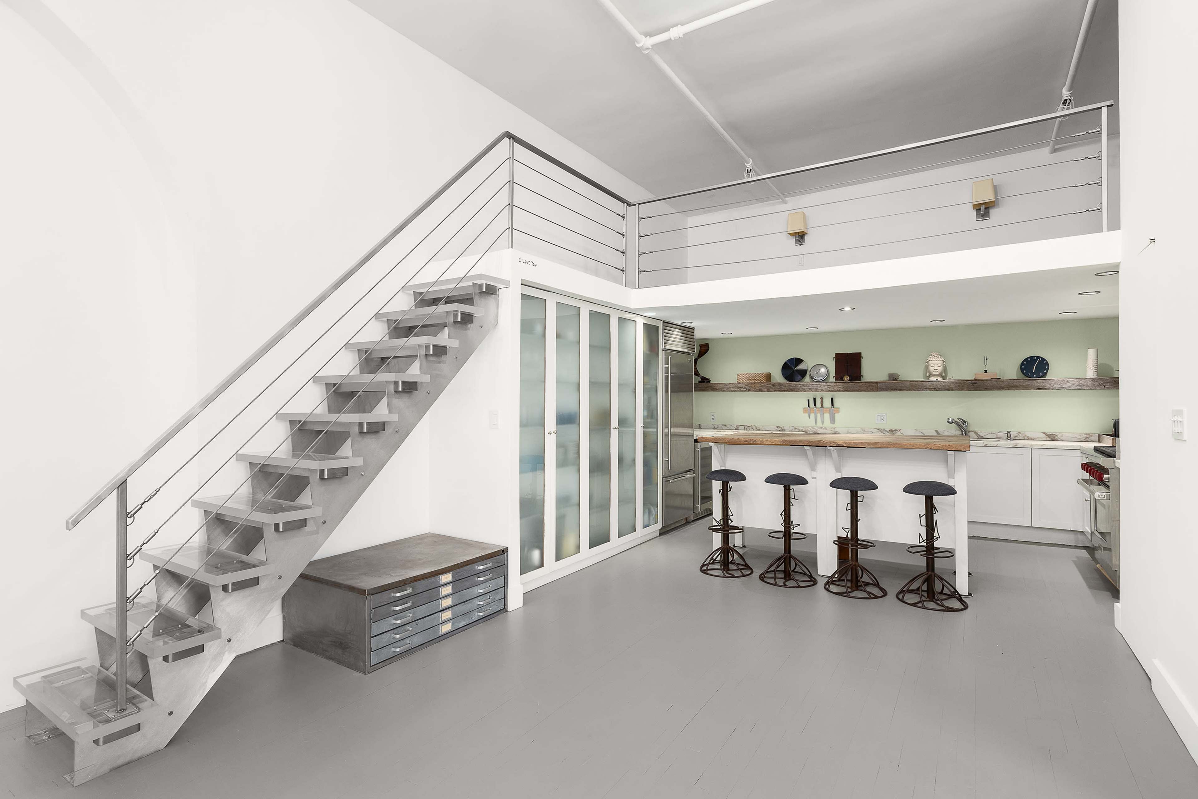 The image shows a modern, minimalist interior featuring a staircase leading to an upper level and a kitchen area with bar stools.