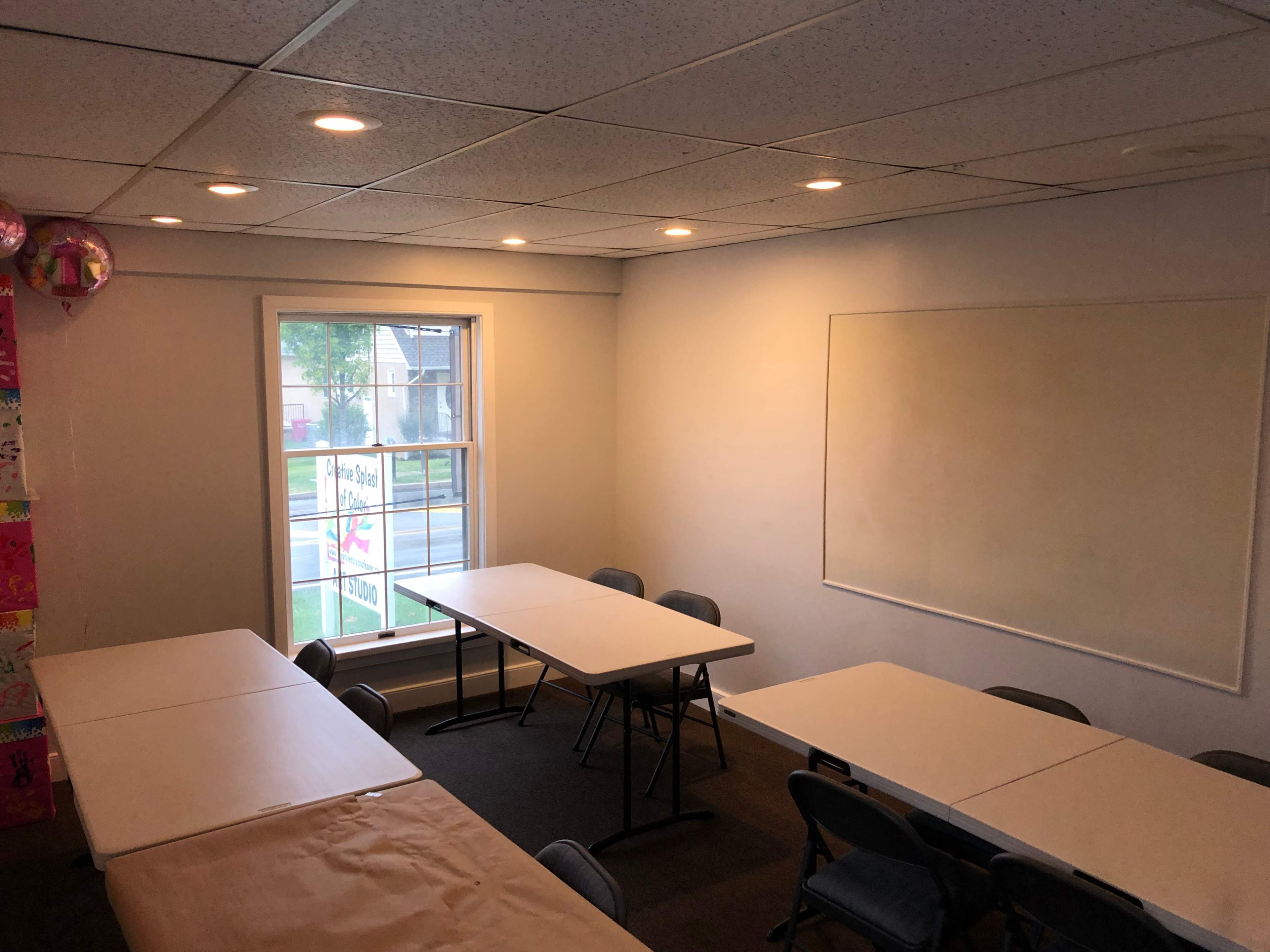 A classroom or meeting room with several tables arranged in a U-shape and a large window letting in natural light.