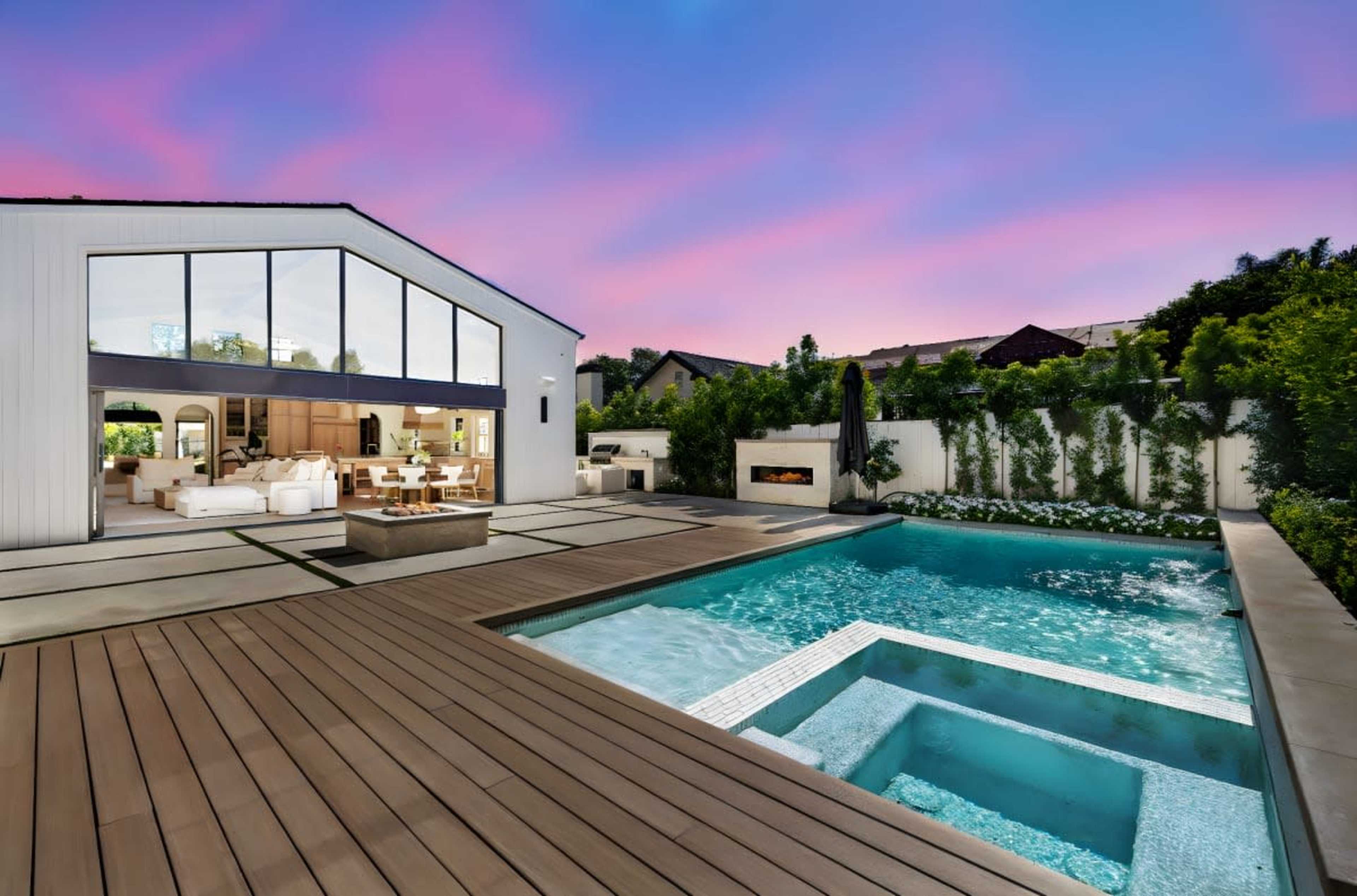 A modern house with large windows overlooks a swimming pool and spa, surrounded by landscaped greenery at dusk.