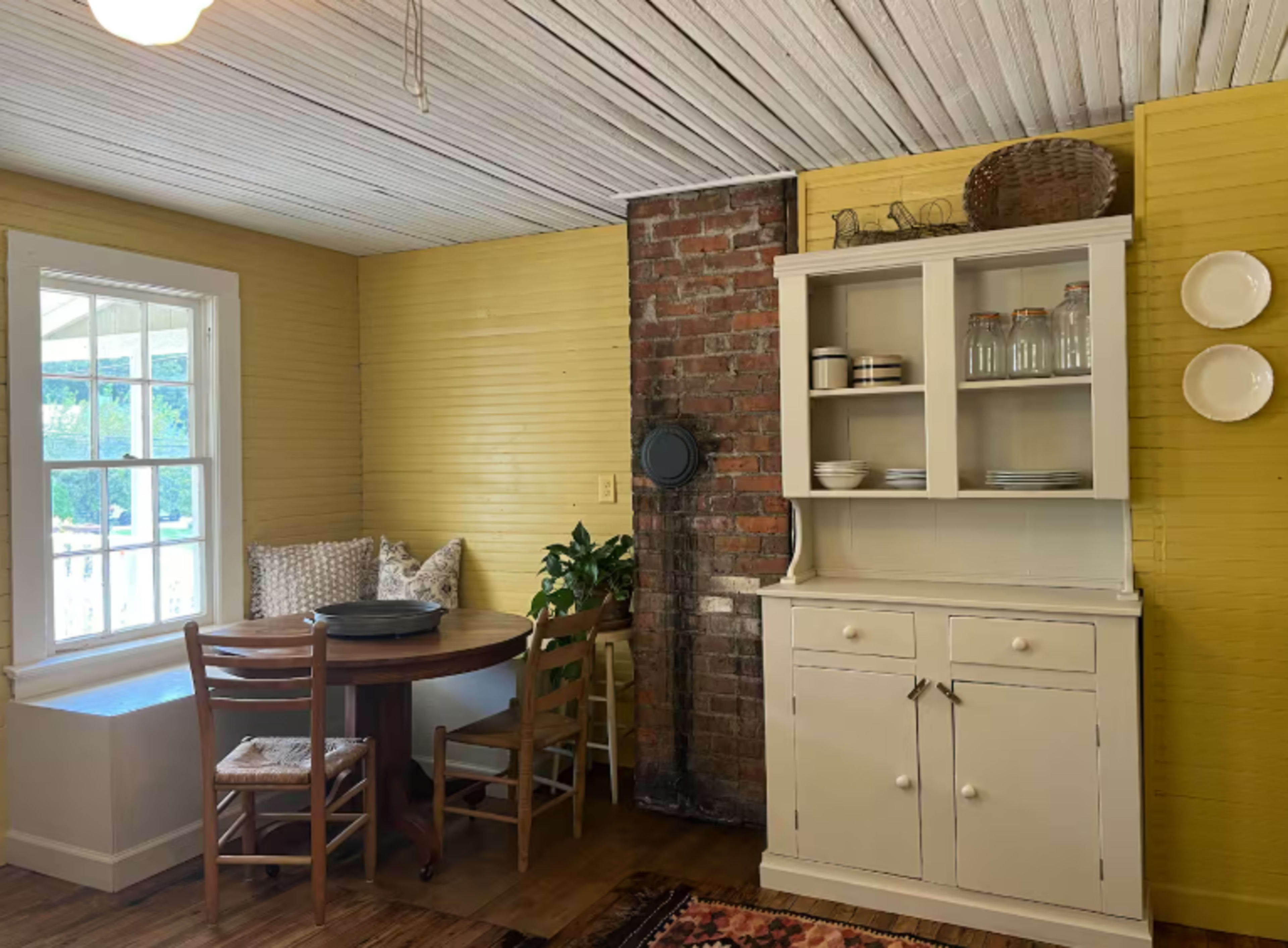 The image shows a cozy dining area with a round wooden table, chairs, a brick wall, and a white cabinet displaying glassware.