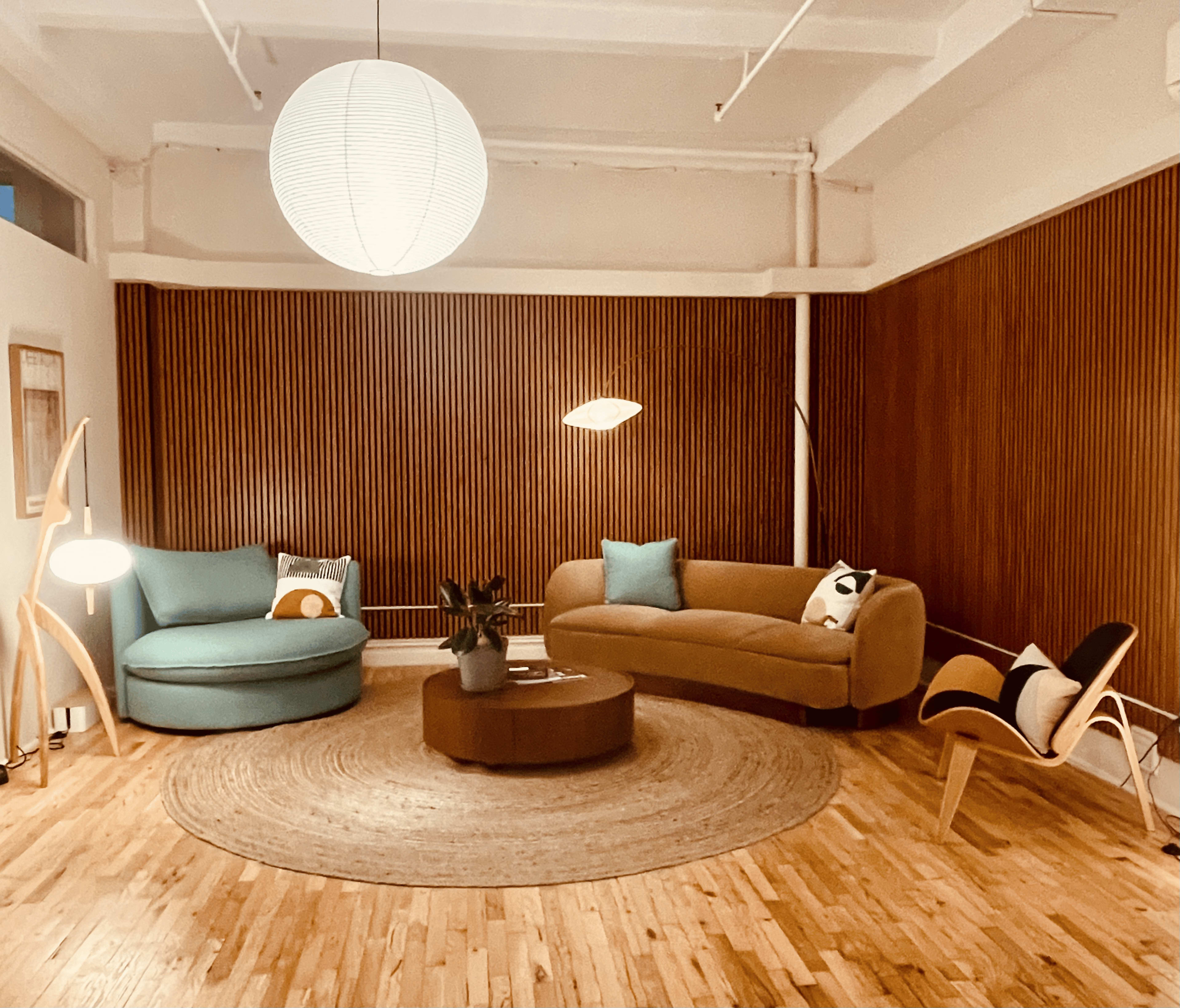 The image shows a modern living room featuring a round area rug, a circular coffee table, and various upholstered seating options against a wood-paneled wall.