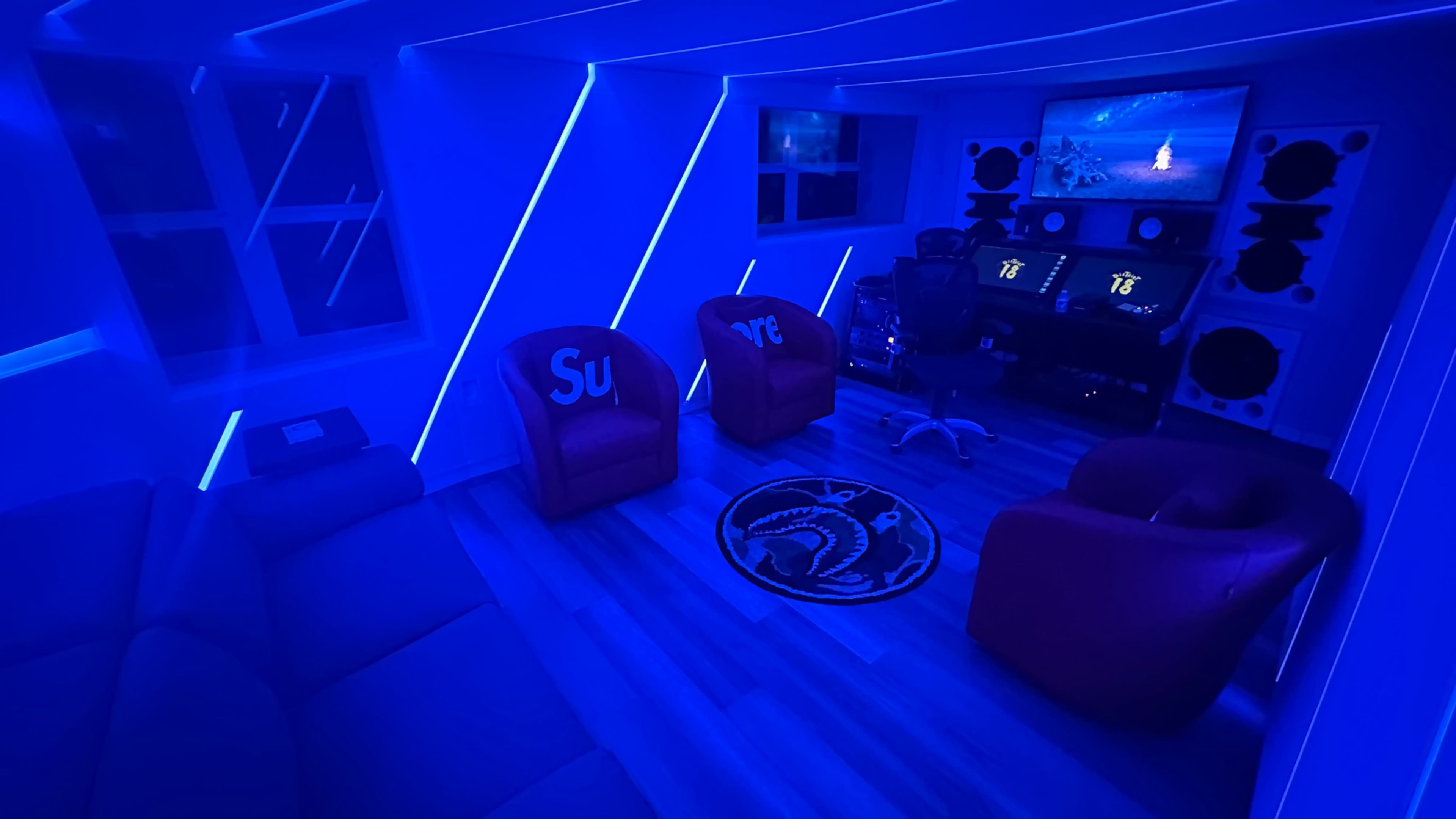 A modern room is illuminated with blue LED lights, featuring two red chairs and a black gaming setup with a large screen and speakers.