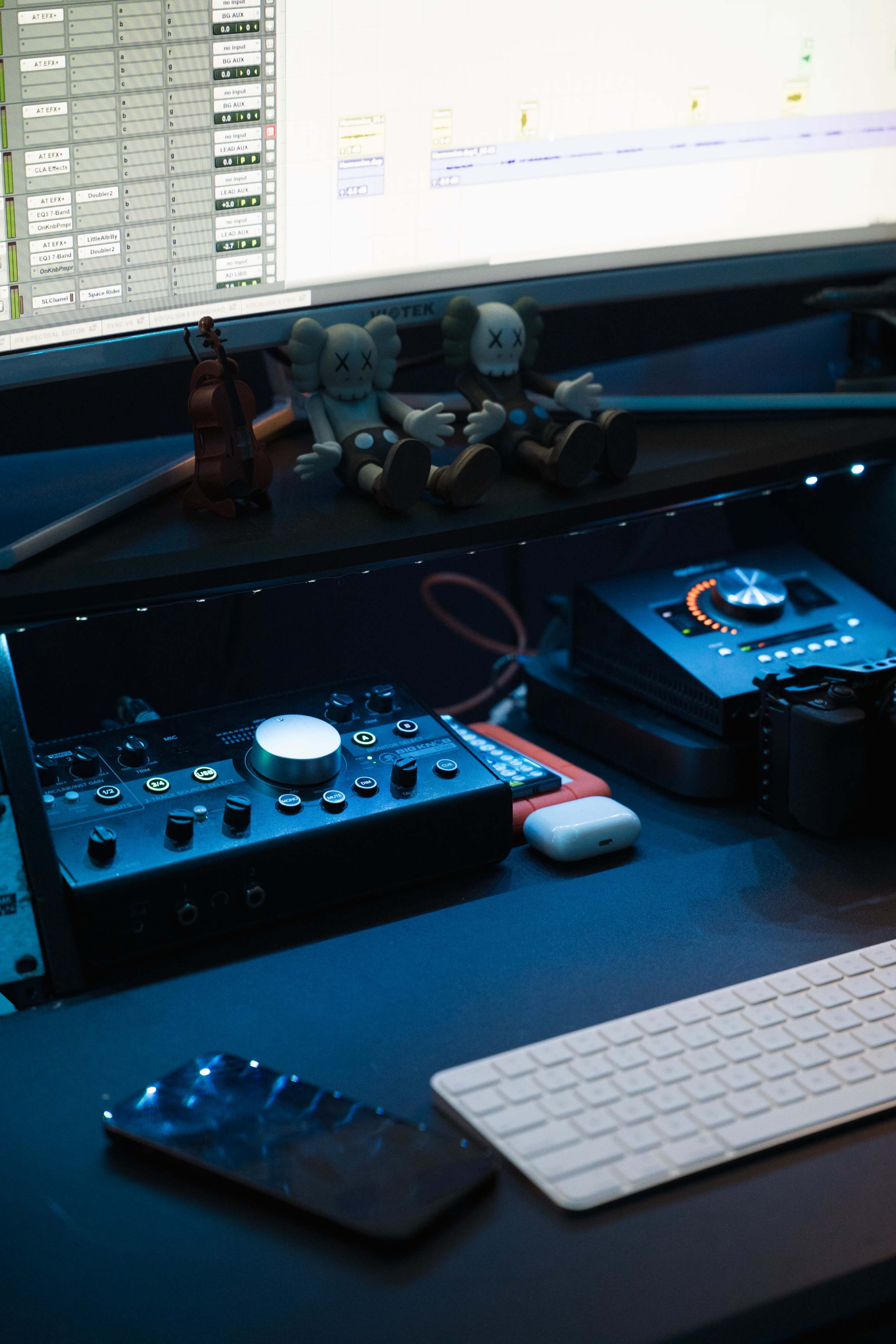 The image shows a music production setup with various equipment, including an audio interface, a keyboard, and figurines, all situated beneath a computer monitor displaying audio tracks.