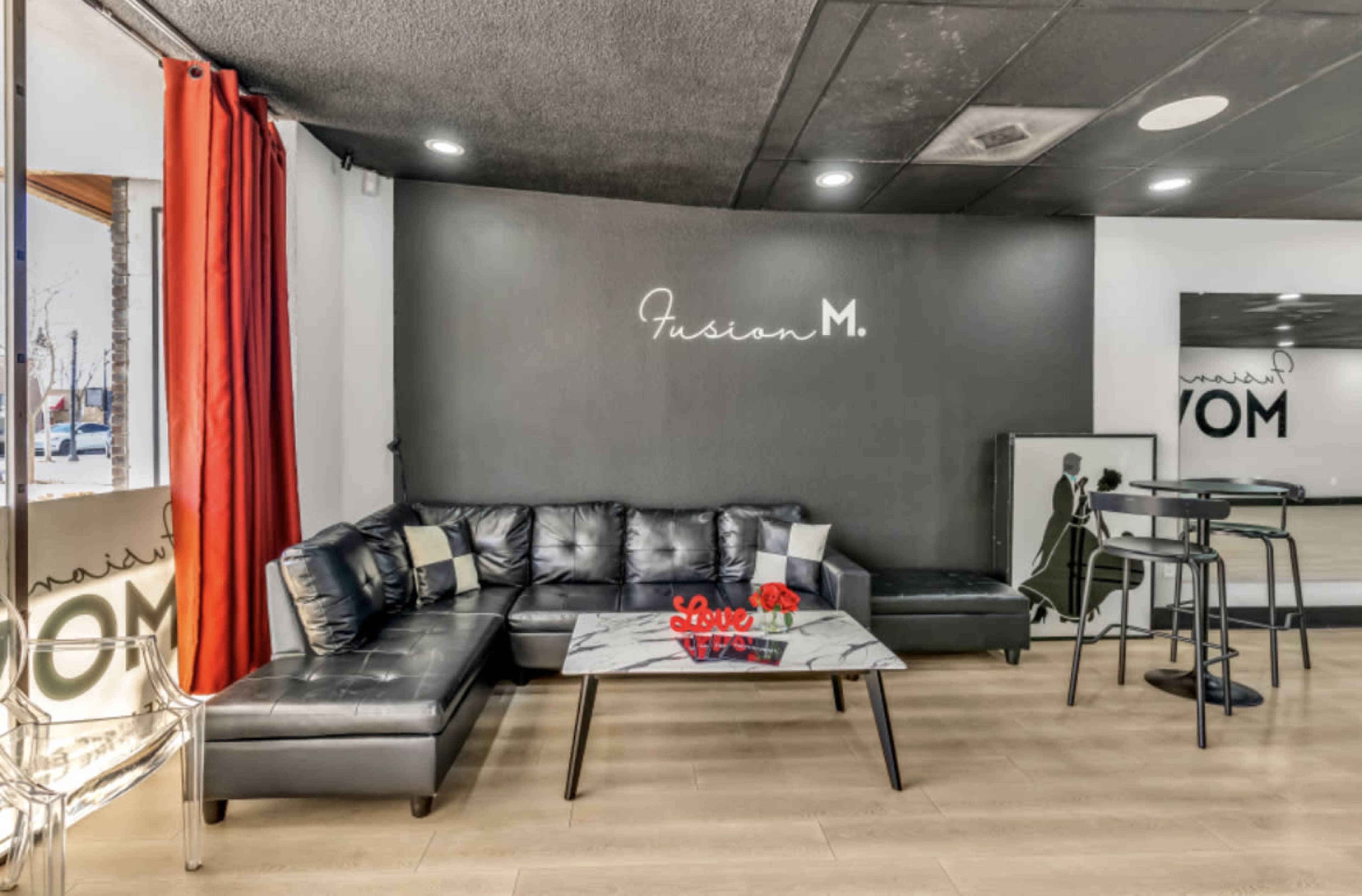 The image shows a modern lounge area featuring a black sectional sofa, a glass coffee table with decorative items, and a wall adorned with the text "Fusion M."