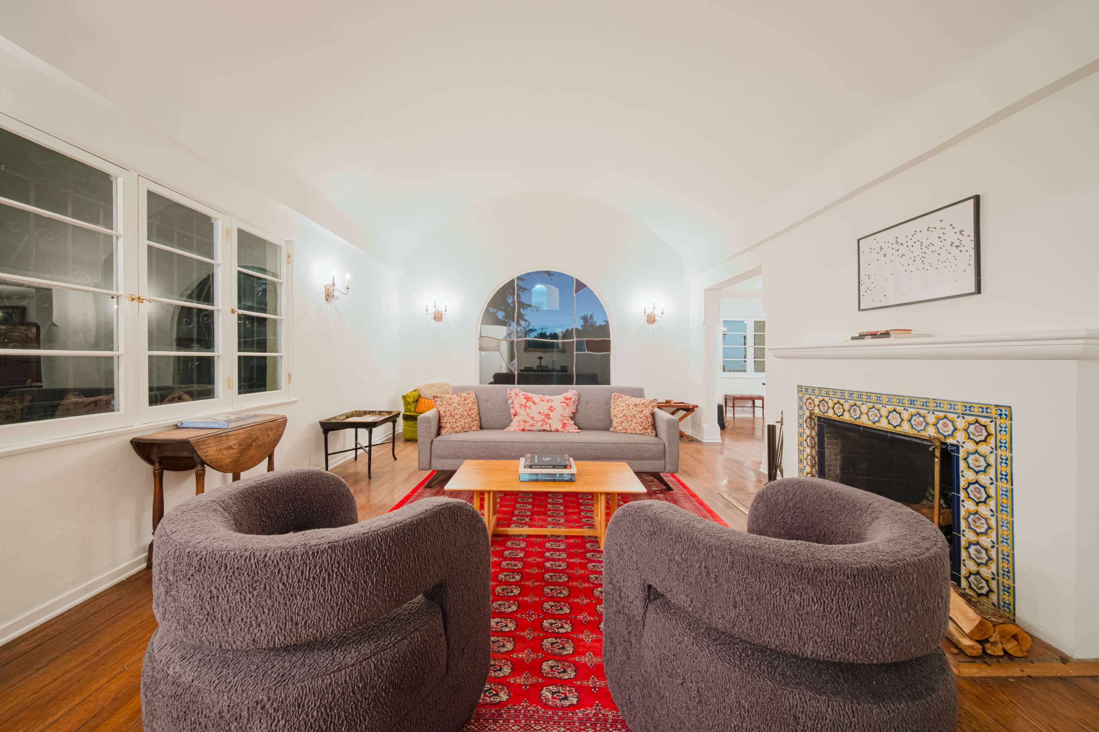 A cozy living room features a gray sofa, round armchairs, a wooden coffee table, and a fireplace, all set on a patterned red rug.