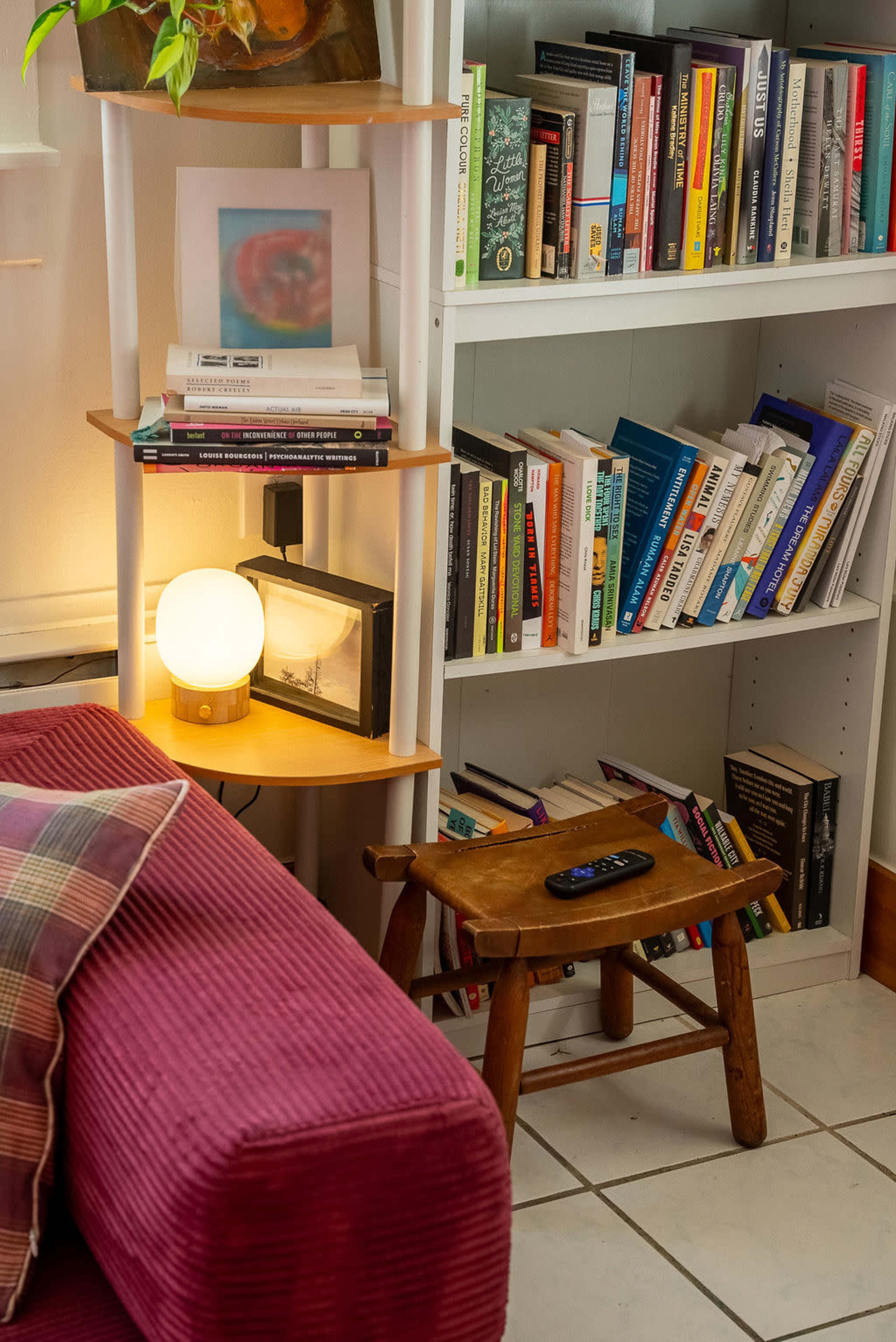 A cozy reading nook features a pink couch, a warm lamp with a round shade, a small wooden table holding a remote, and a bookshelf filled with various books.
