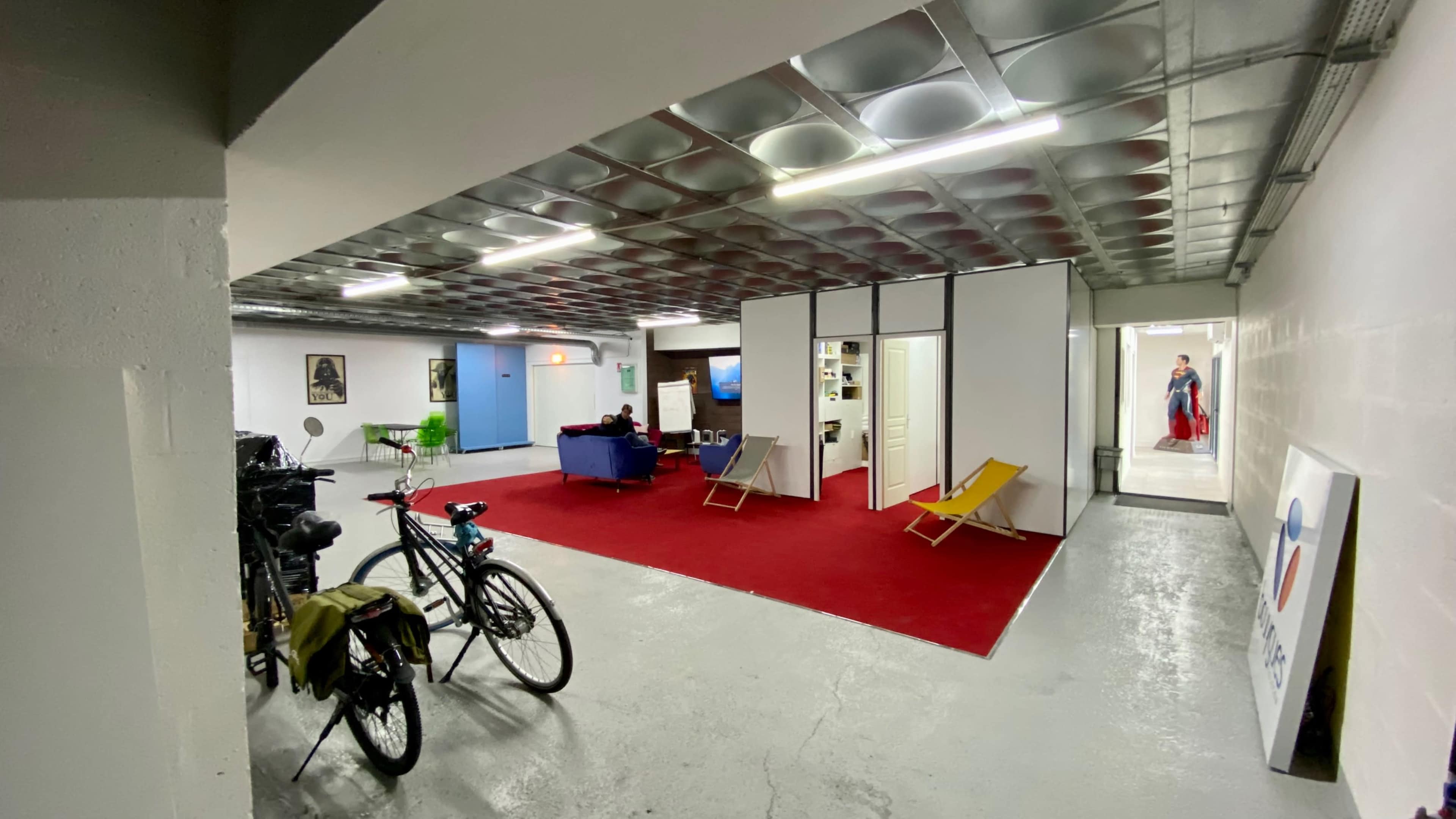A spacious, modern basement lounge area with a red carpet, a blue couch, a small TV area, bicycles parked on one side, and a view of a hallway leading to additional rooms.