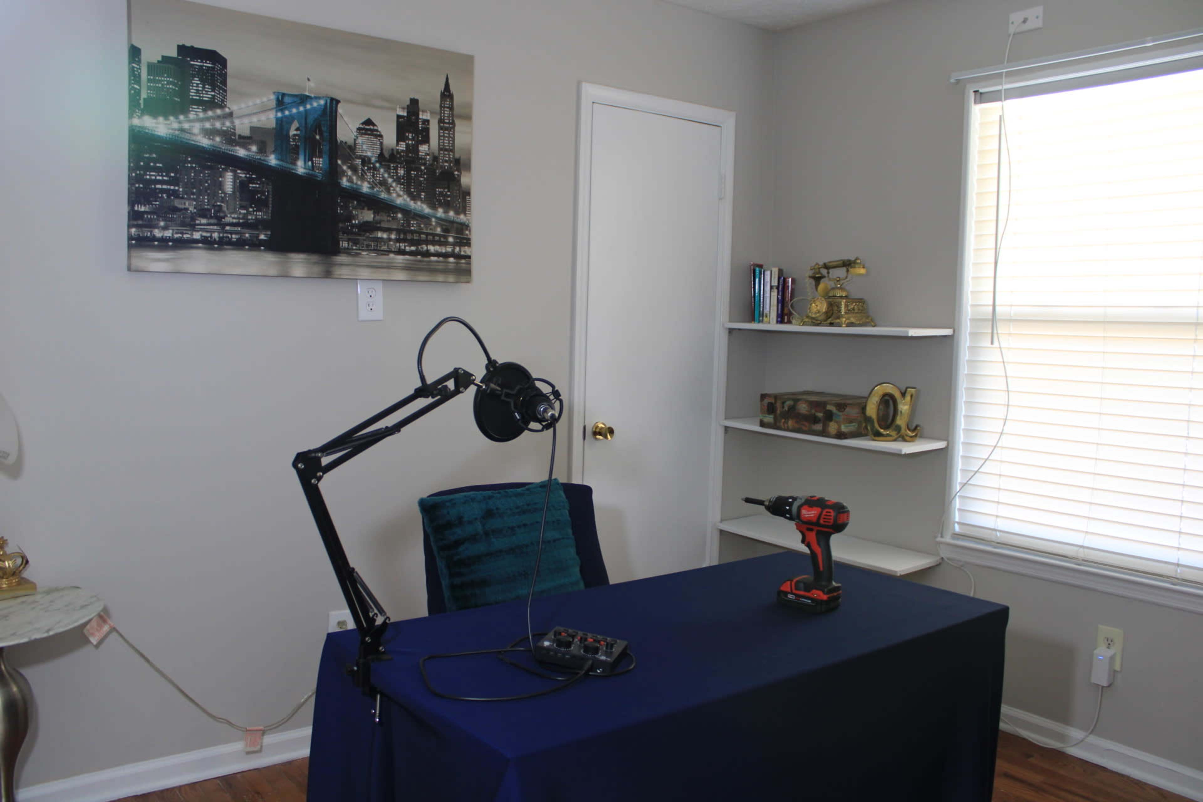 The image shows a small office space with a blue table, a microphone on an arm, a power drill, and a shelf against a light-colored wall decorated with a cityscape picture.