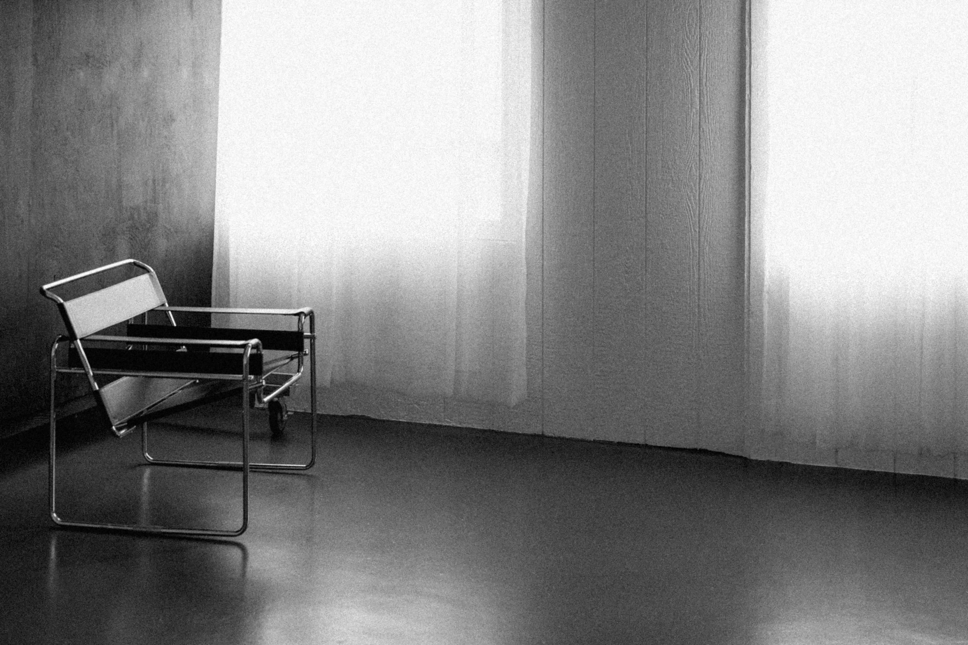 A minimalist, metallic chair sits in a corner beside two large, white-curtained windows.