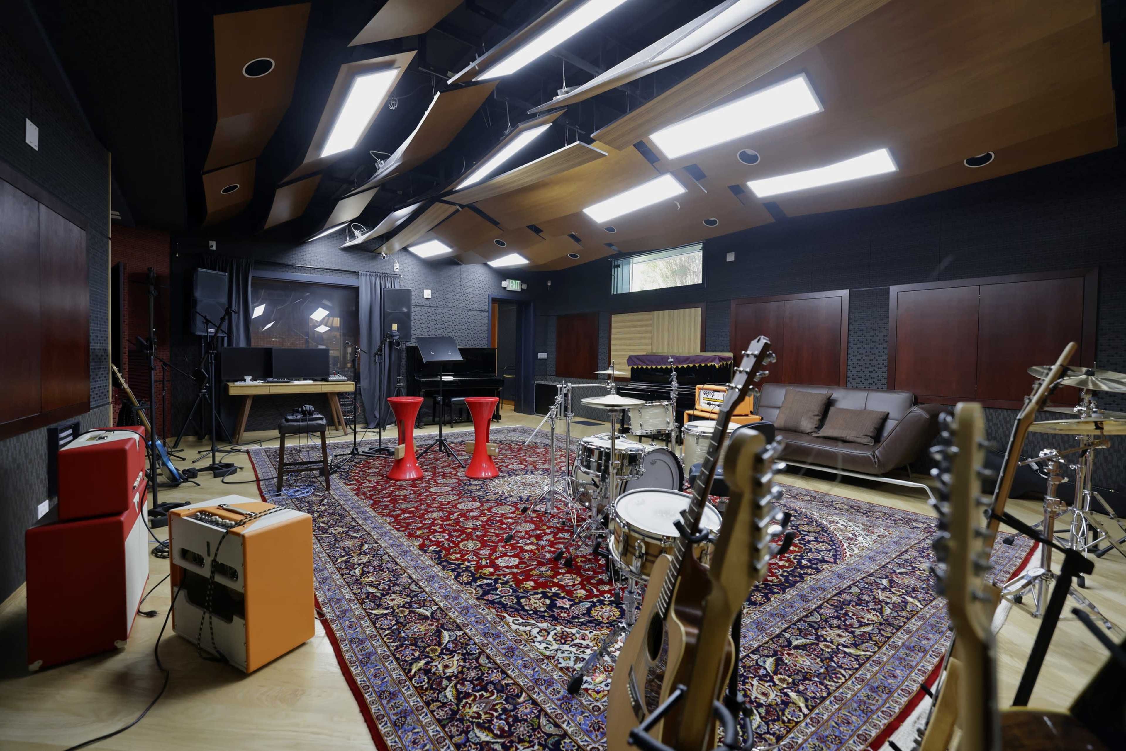 A music studio with various instruments, a sofa, and red stools arranged on a patterned rug.