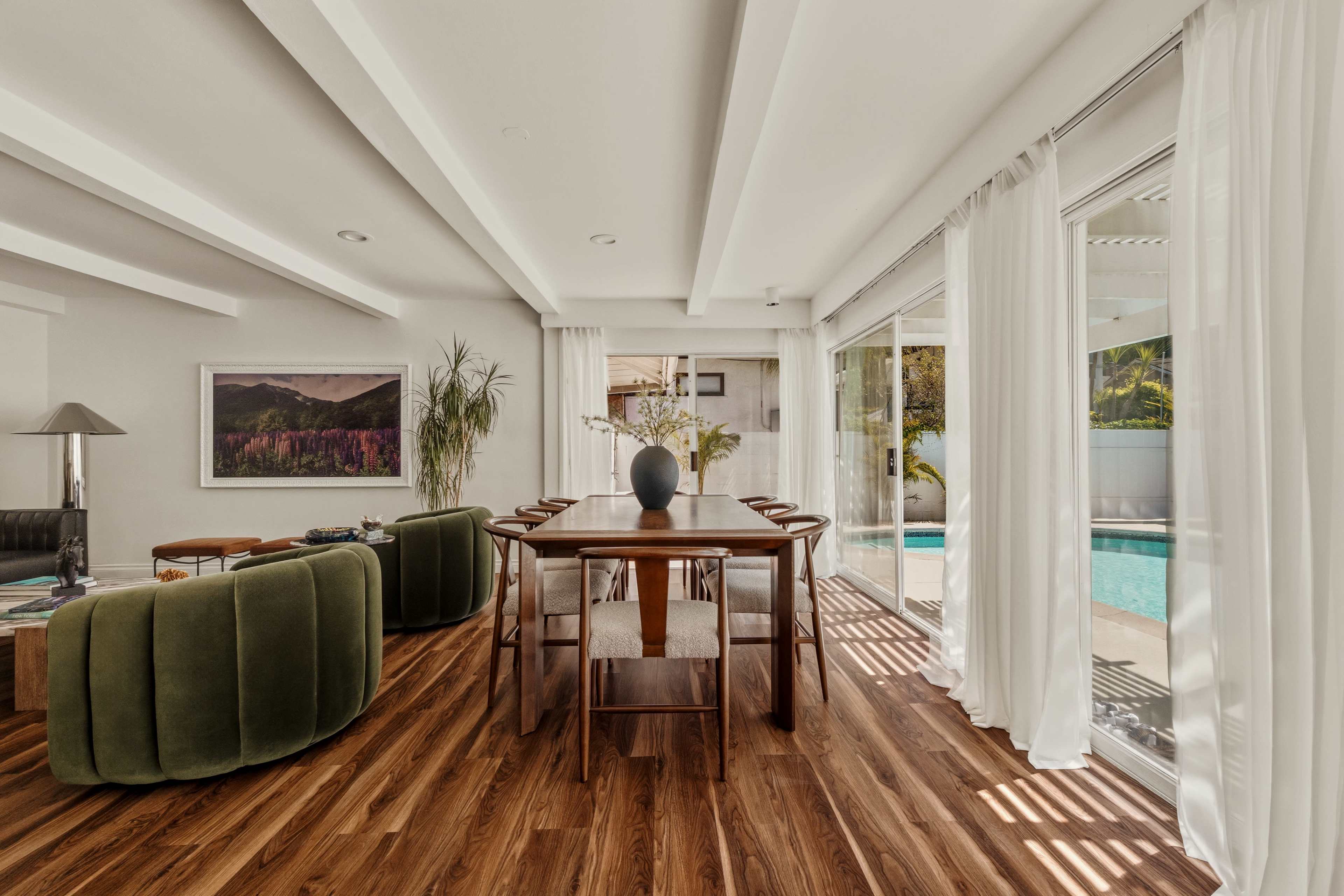 A modern dining area features a wooden table surrounded by chairs, with large windows overlooking a pool and lush outdoor space.