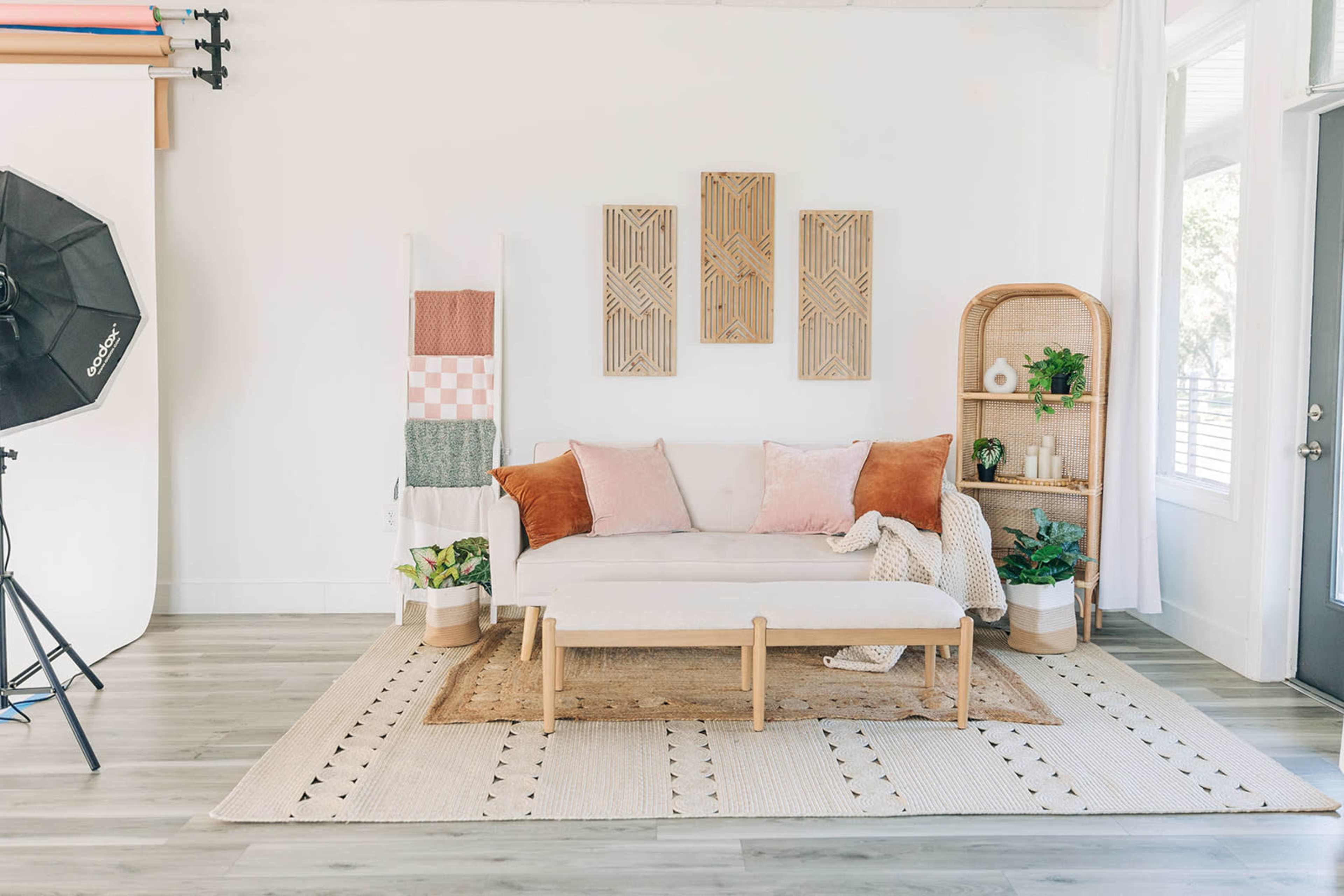 The image shows a modern, bright living room featuring a light-colored sofa with decorative cushions, a wooden coffee table, and various indoor plants arranged on a shelf and around the room.