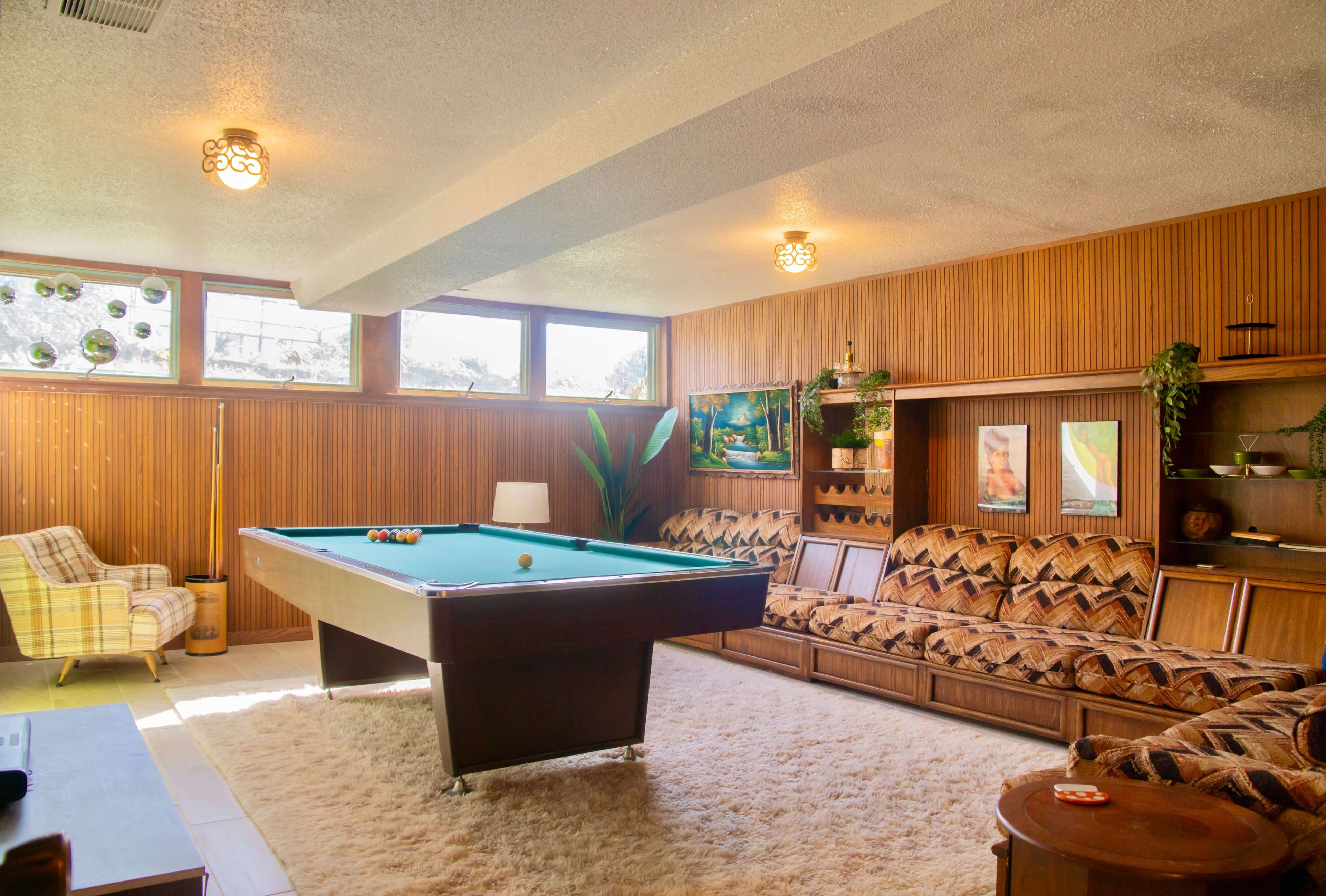The image shows a retro game room with a green pool table, patterned brown sofas, and wooden paneling on the walls.