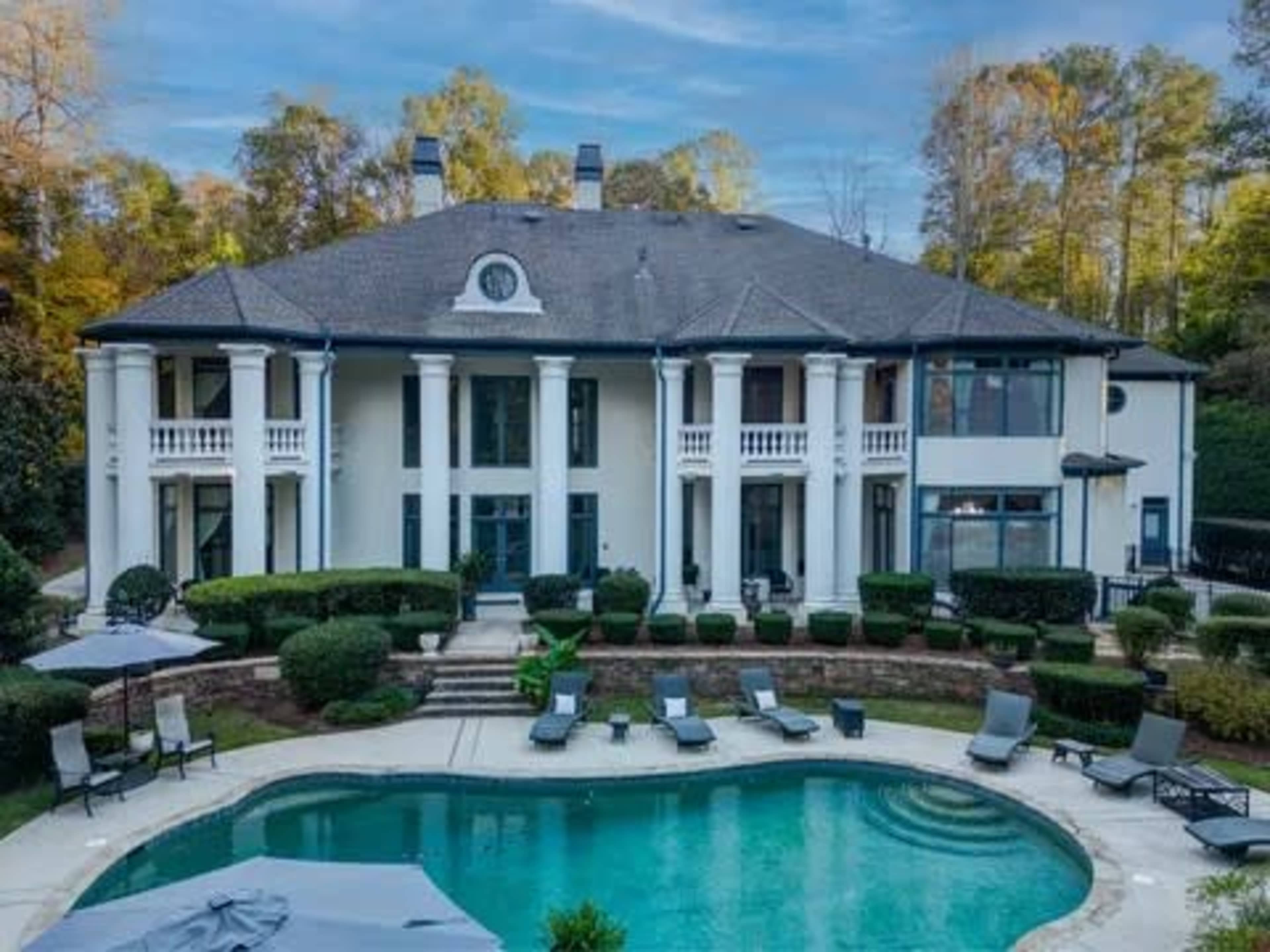 The image shows a large white mansion with a swimming pool in the foreground, surrounded by neatly trimmed bushes and deck chairs.