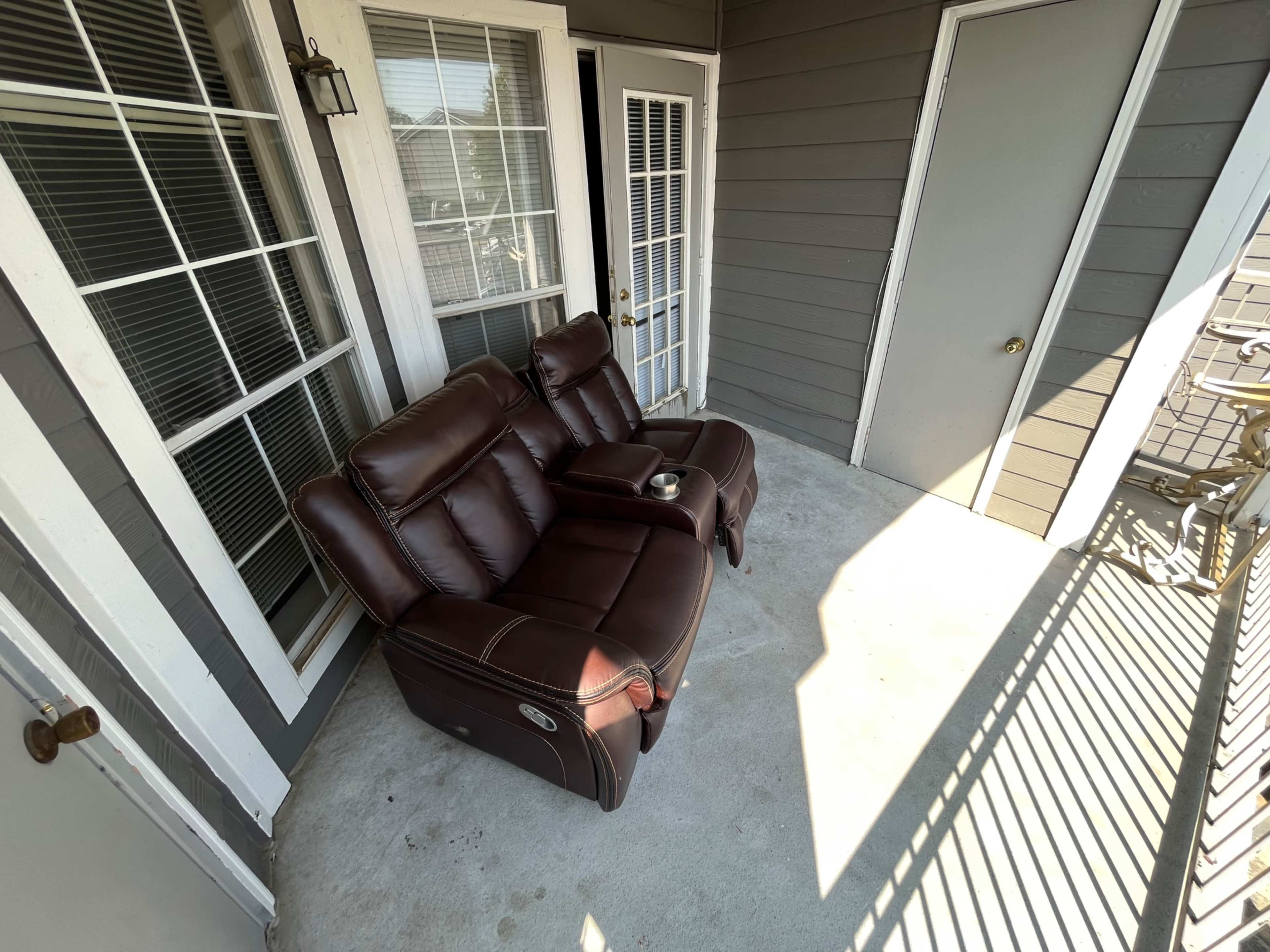 A reclining loveseat with a cup holder sits on a concrete porch next to a door and windows.