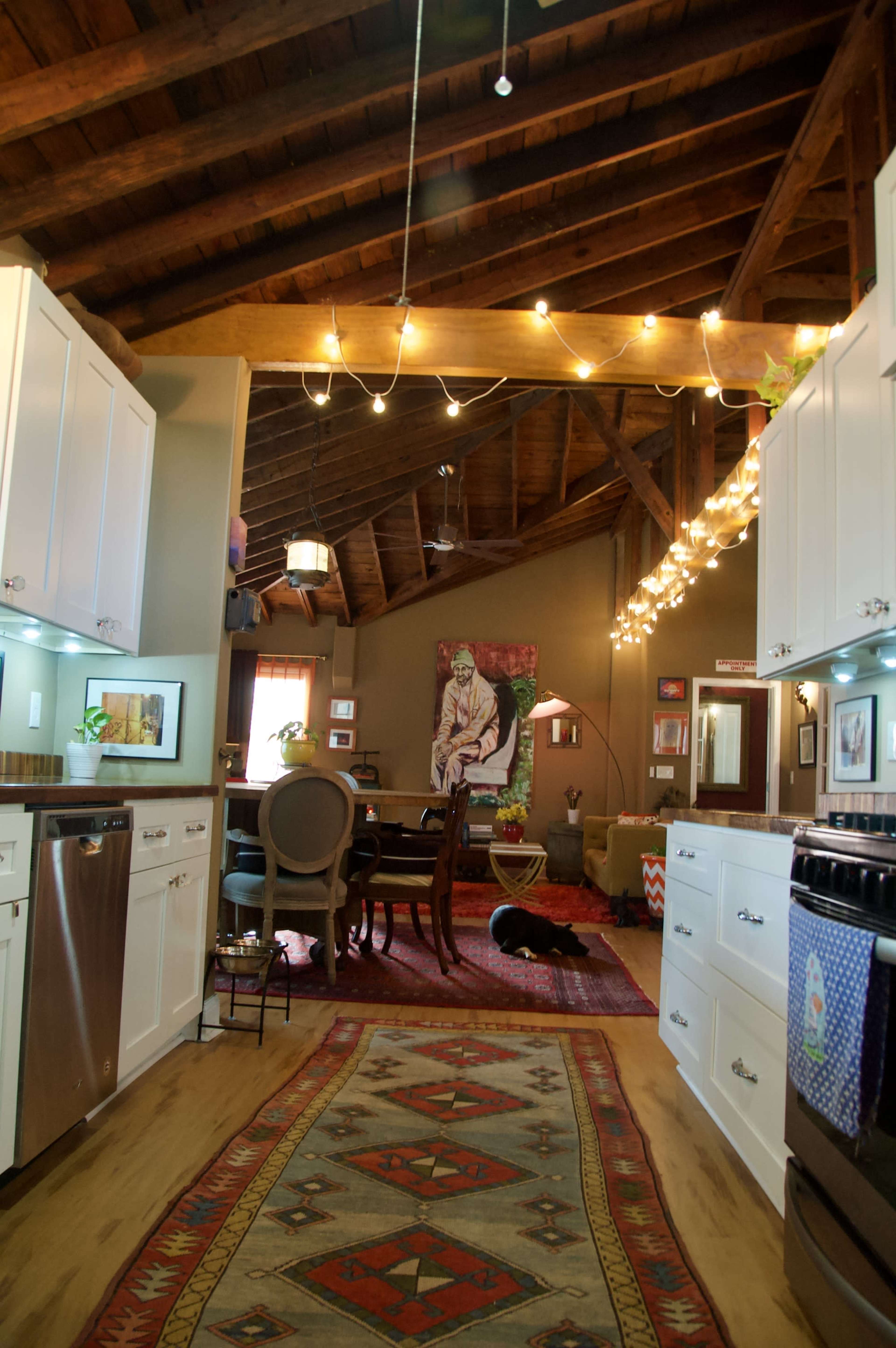 The image shows a spacious kitchen and dining area featuring wooden beams, white cabinetry, a large rug, and a pet dog lying on the floor.