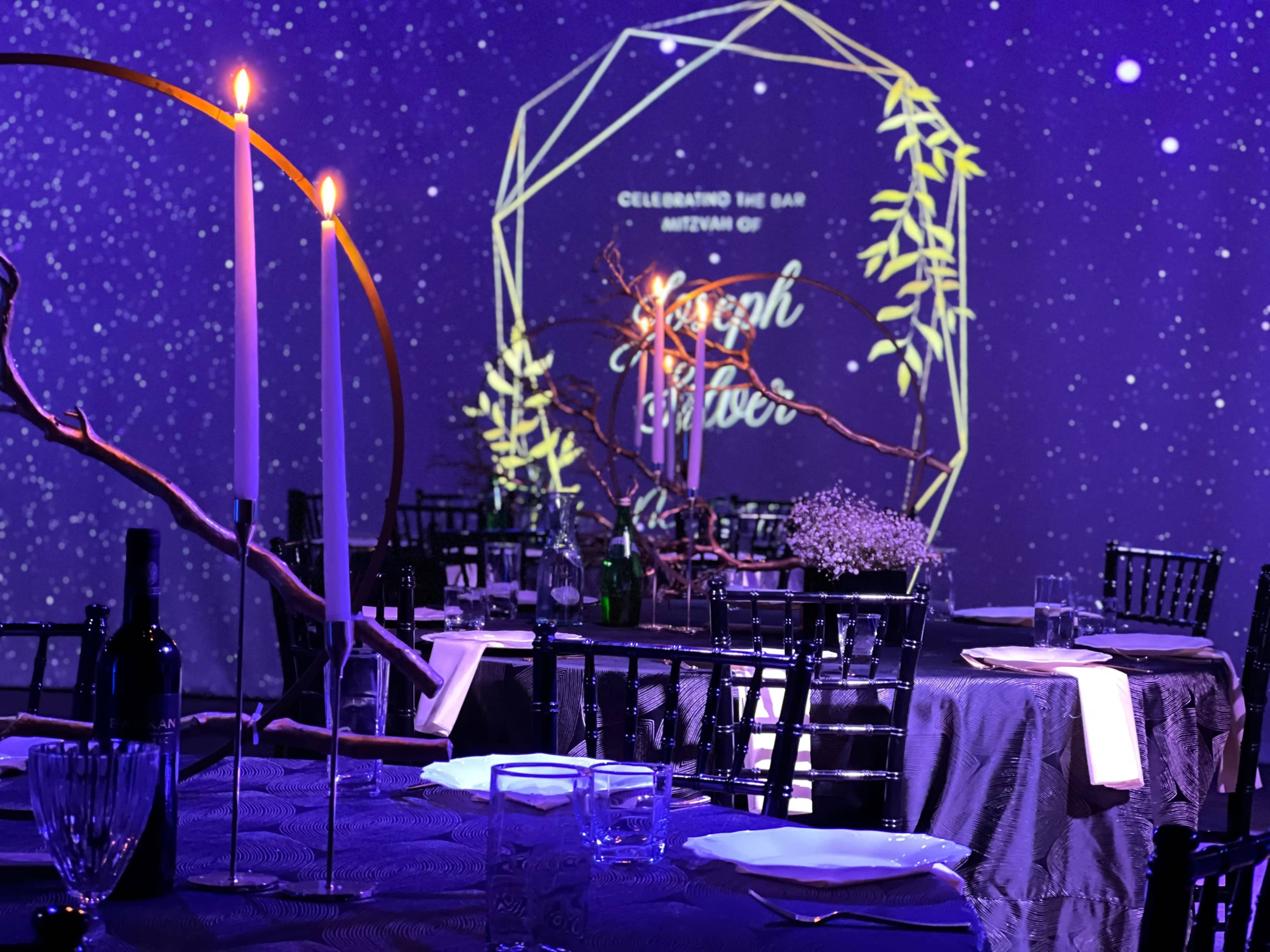 A elegantly set banquet table features candles and floral arrangements, with a backdrop displaying a starry theme and the name "Joseph" illuminated.