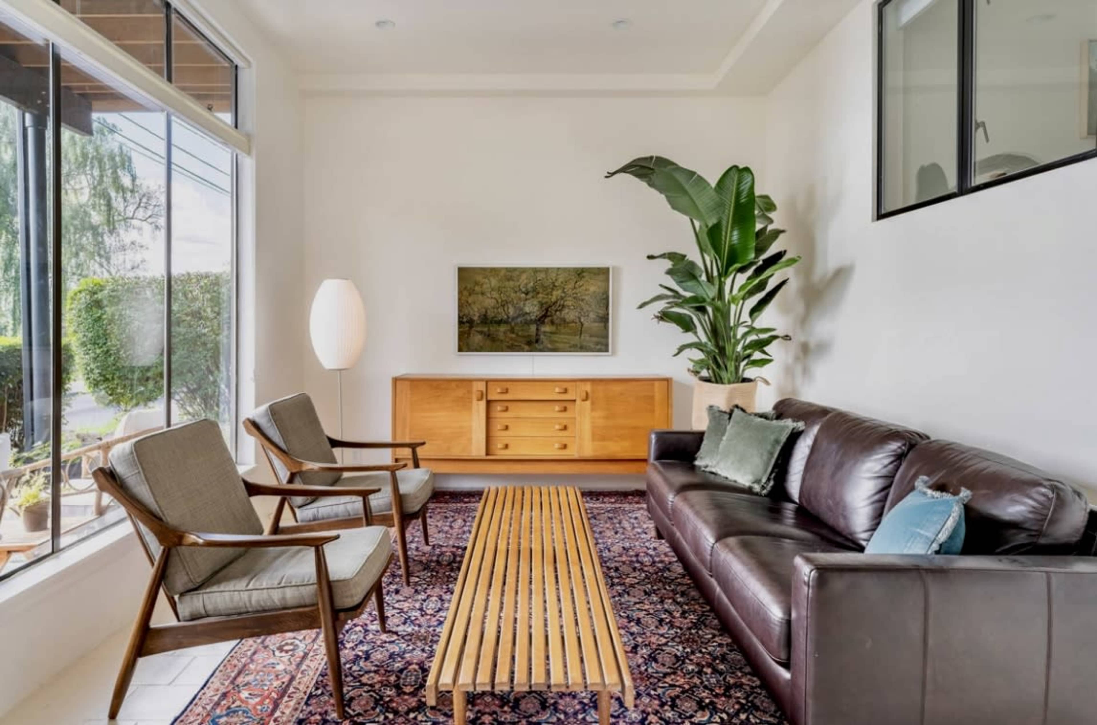 A well-lit living room features a leather sofa, two wooden chairs, a striped coffee table, and a large plant beside a cabinet and an artwork on the wall.