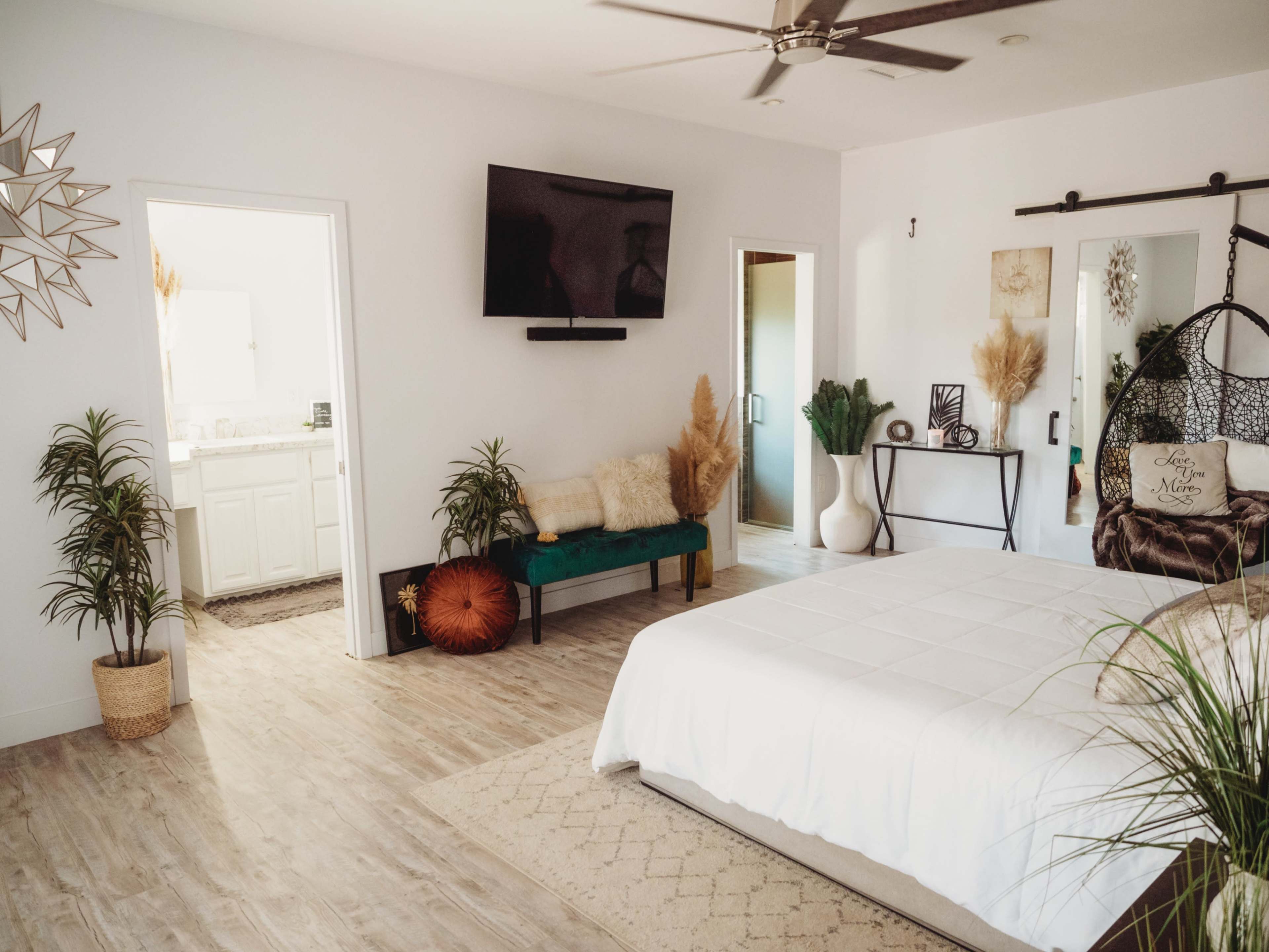 A spacious bedroom featuring a white bed, a green bench, a television mounted on the wall, and a bathroom accessible through an adjoining door.