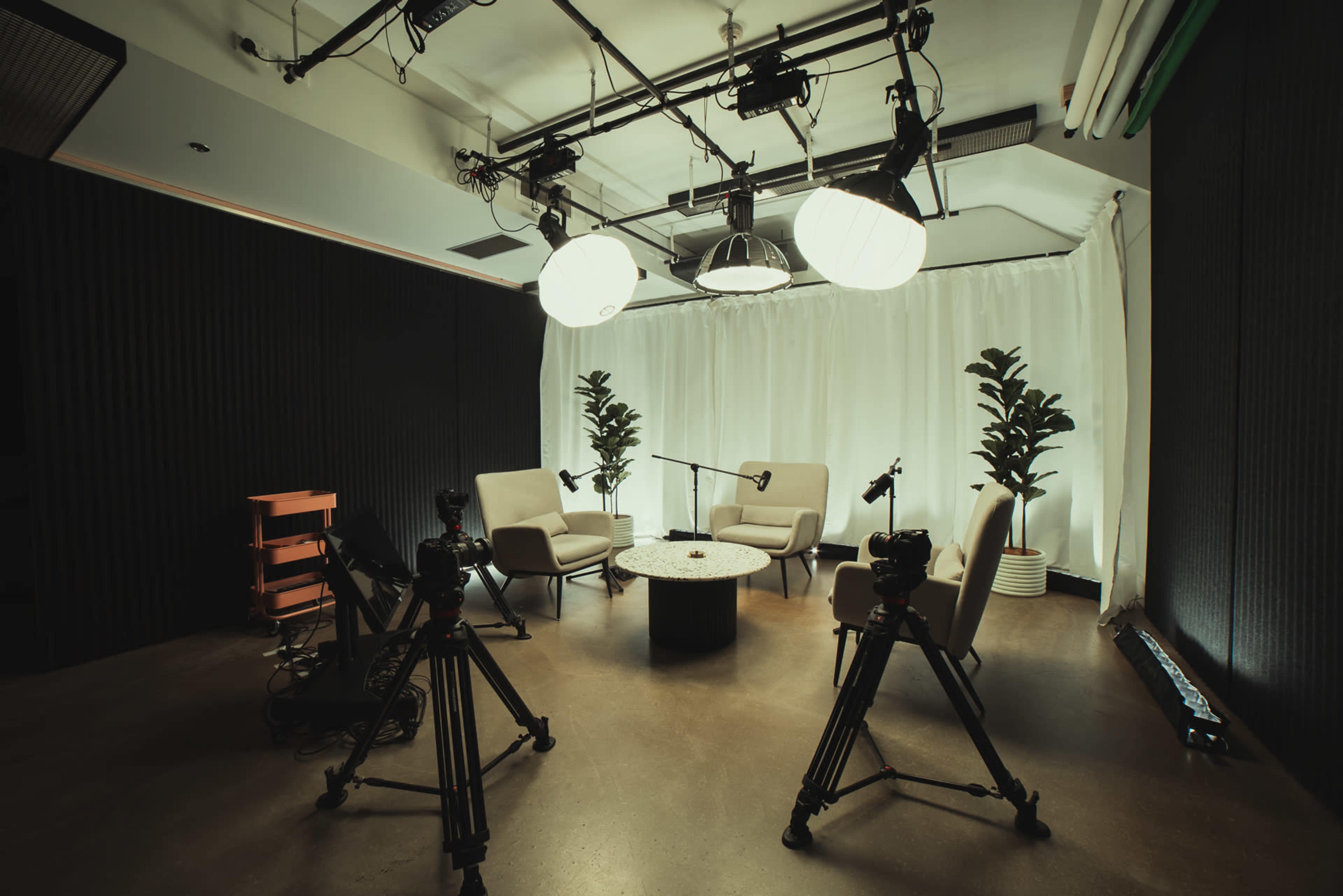 A modern studio setup featuring two chairs, a round table, and several cameras on tripods, with soft lighting and decorative plants.