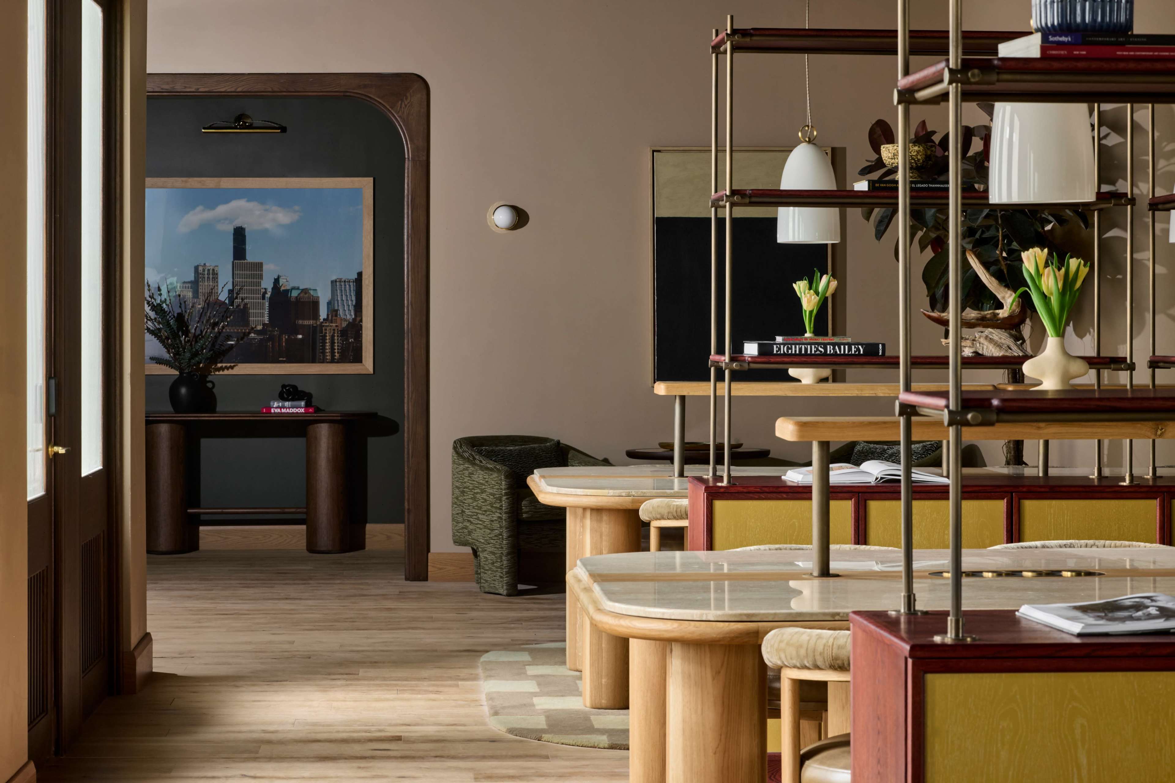 The image shows a modern interior of a restaurant or café with wooden flooring, stylish furniture, and decorative plants, featuring an artwork on the wall.