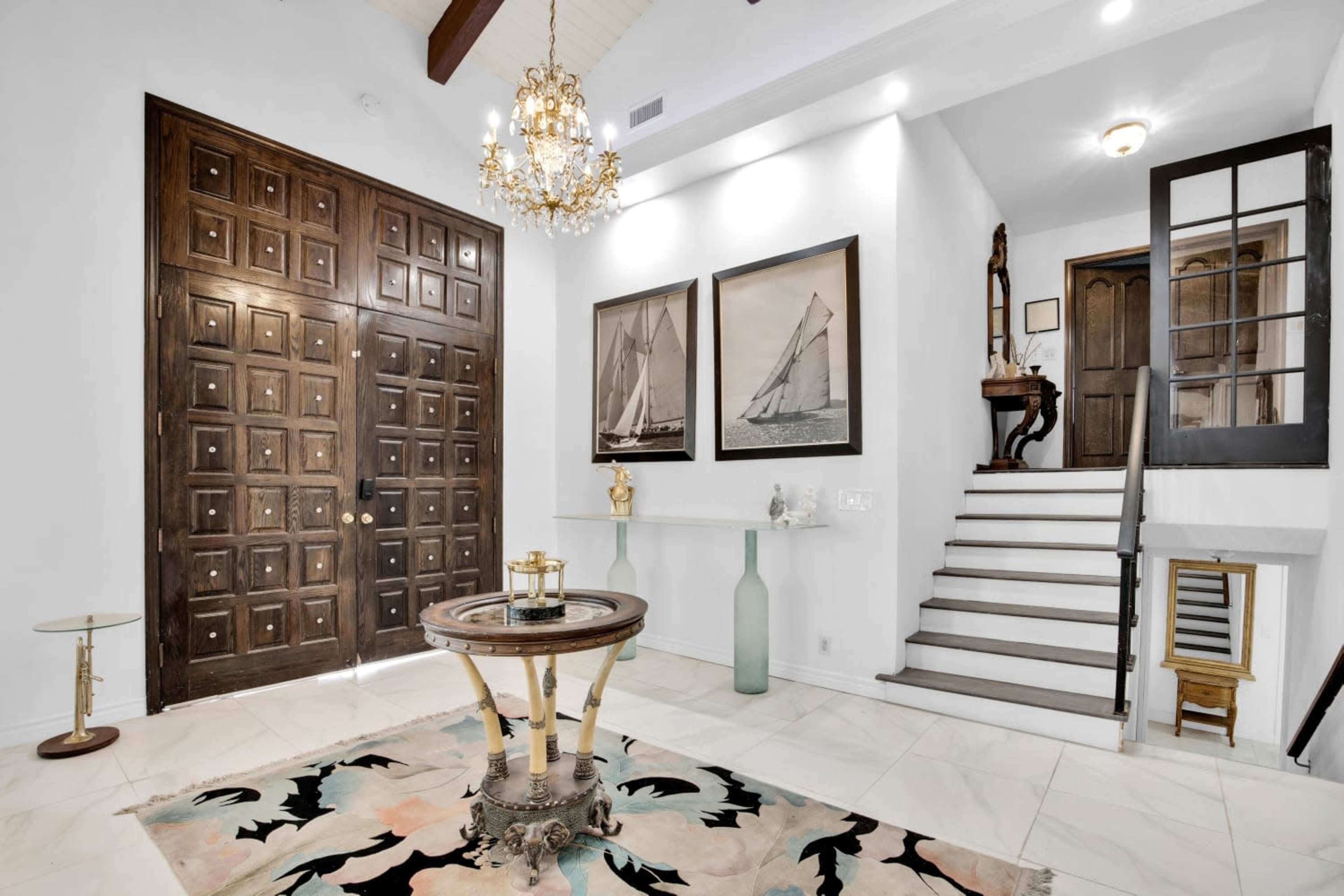 A spacious entryway featuring a large wooden door, decorative chandeliers, framed pictures of sailboats, and a staircase leading to an upper level.