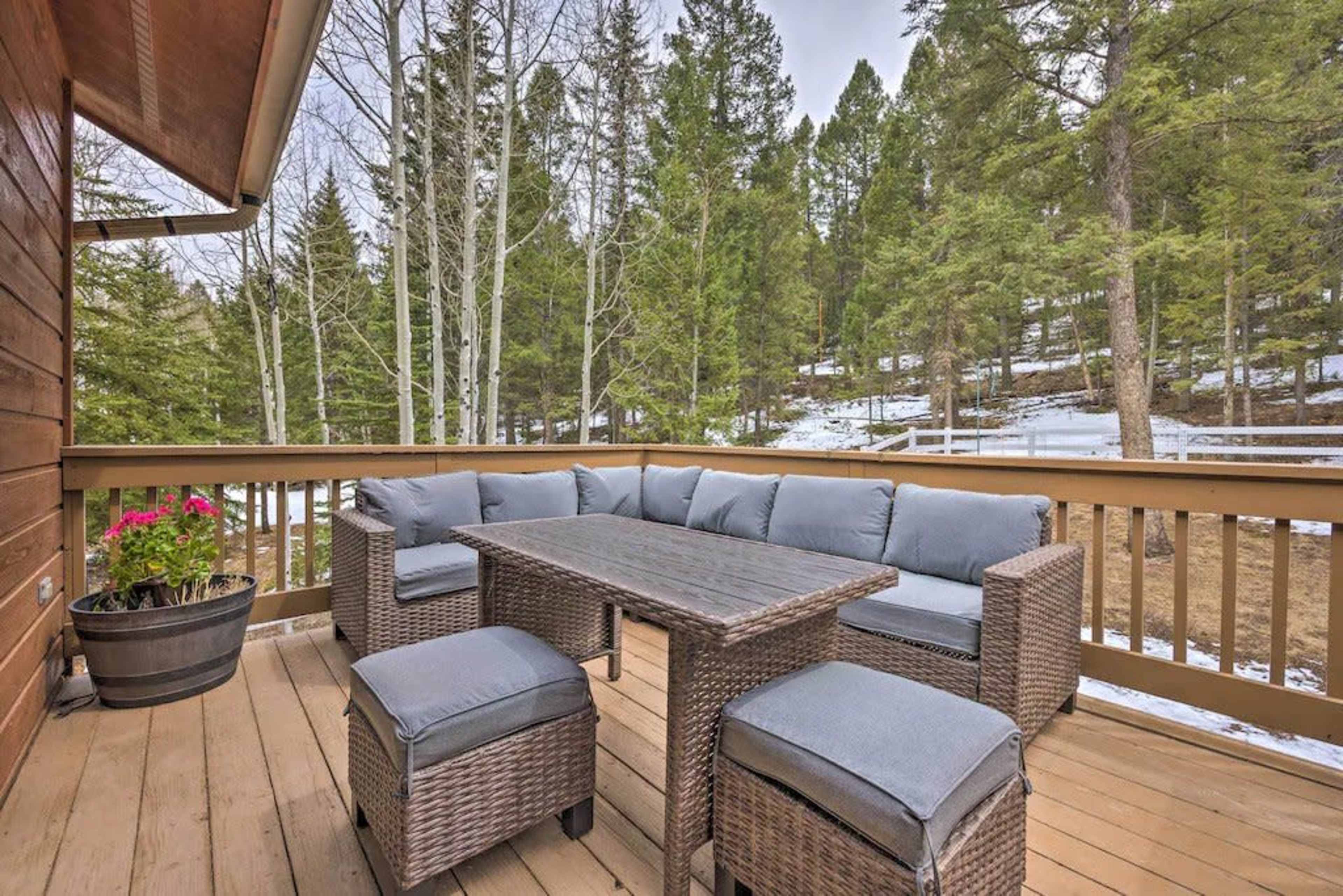 A wooden deck features a rectangular table surrounded by cushioned seating, with planters and a backdrop of tall pine trees and a hint of snow.