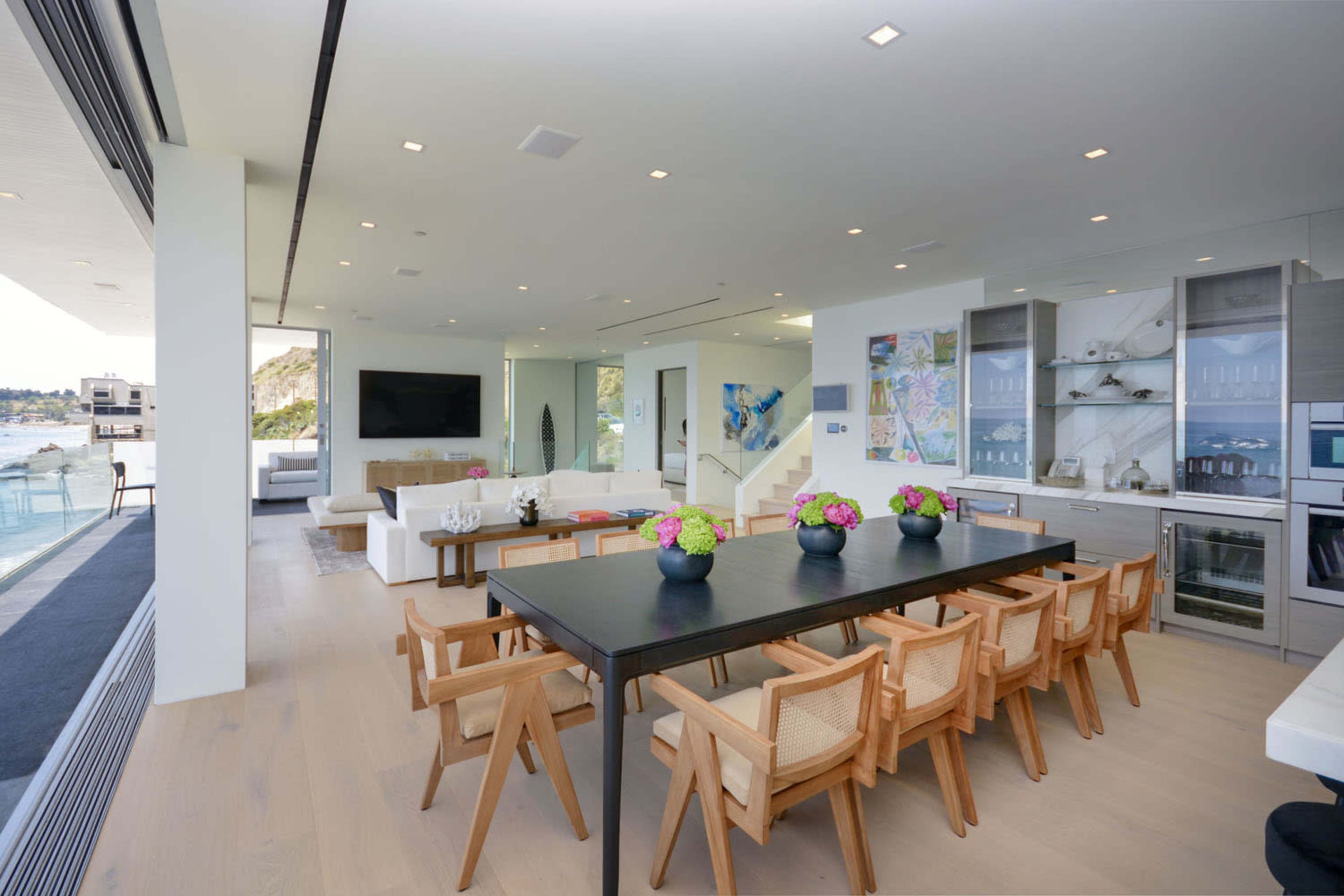 A modern dining area with a long table surrounded by wooden chairs, adjacent to a sleek kitchen and a living space with large windows overlooking a view.