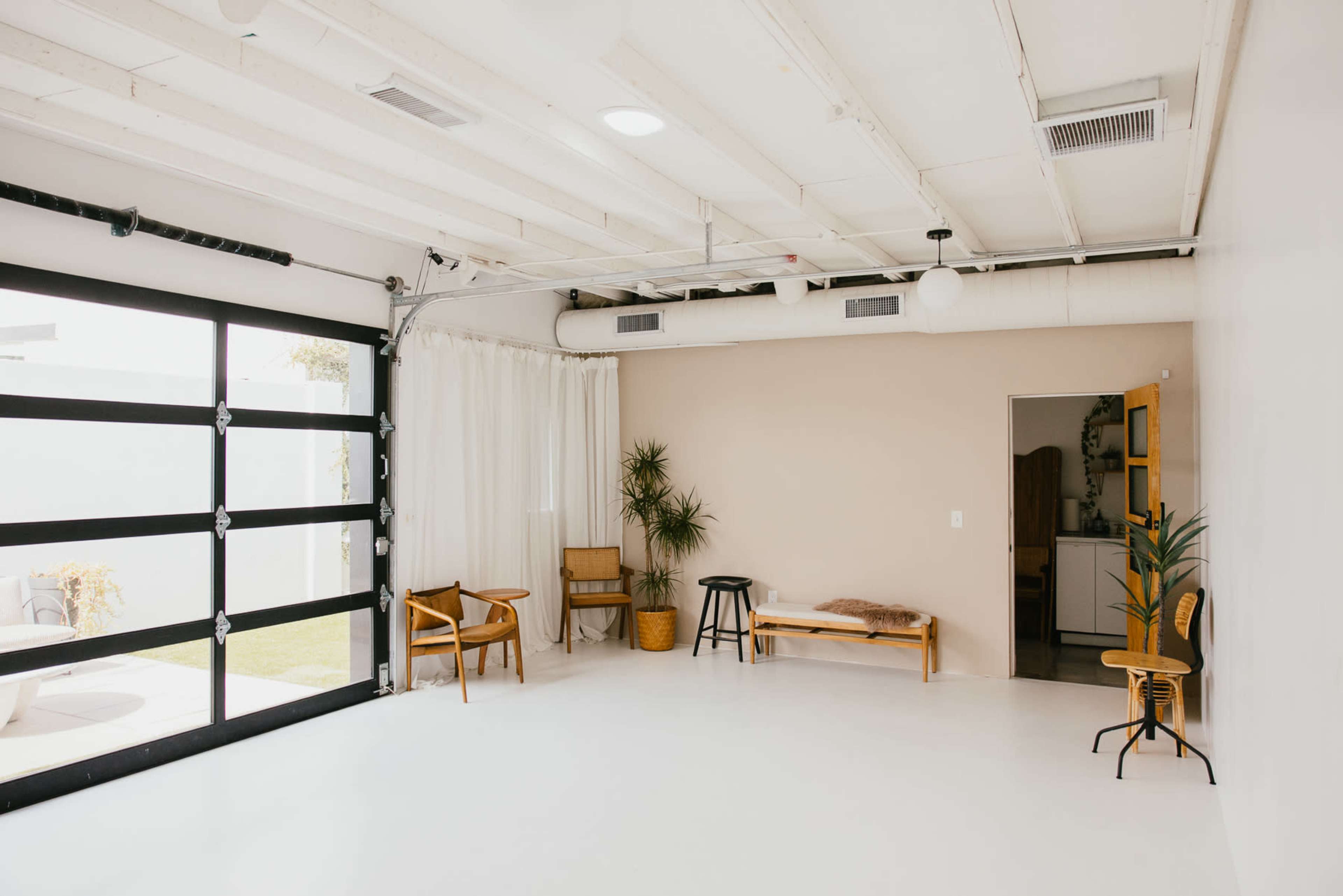 The image shows a minimalist indoor space with white walls, a concrete floor, and a large garage door opening to natural light, furnished with a few chairs, a bench, and a plant.